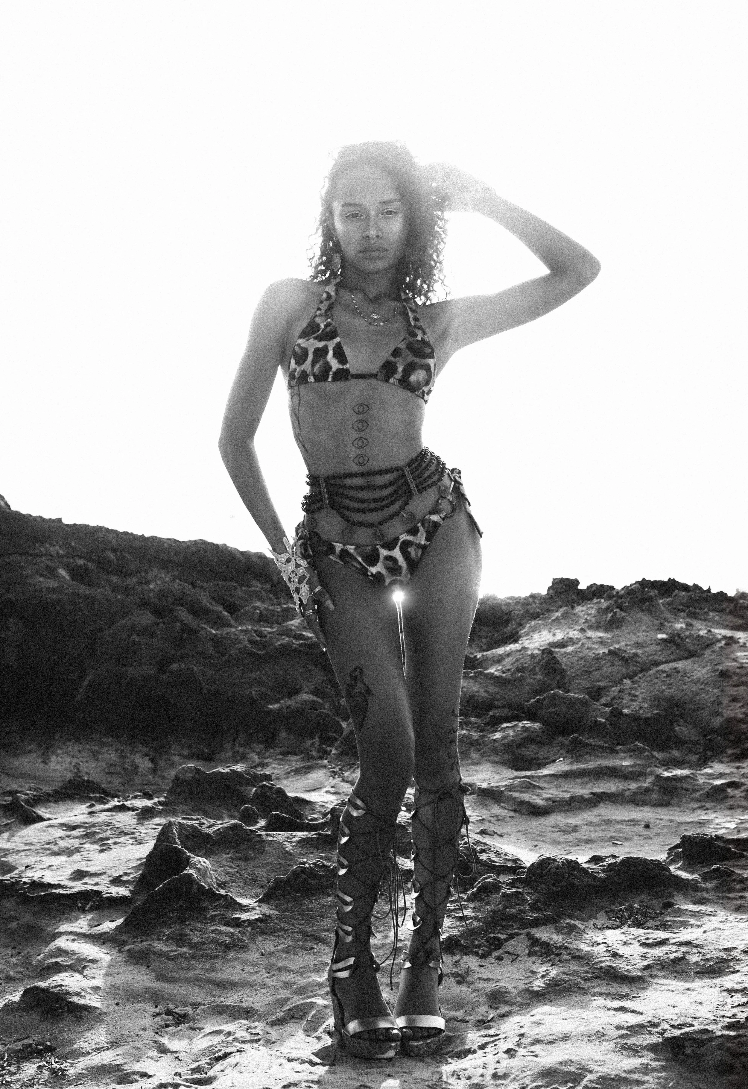 A woman in a leopard print bikini, standing on rocky terrain, wearing lace-up high-heeled sandals, and accessories, with the sun behind her creating a silhouette.