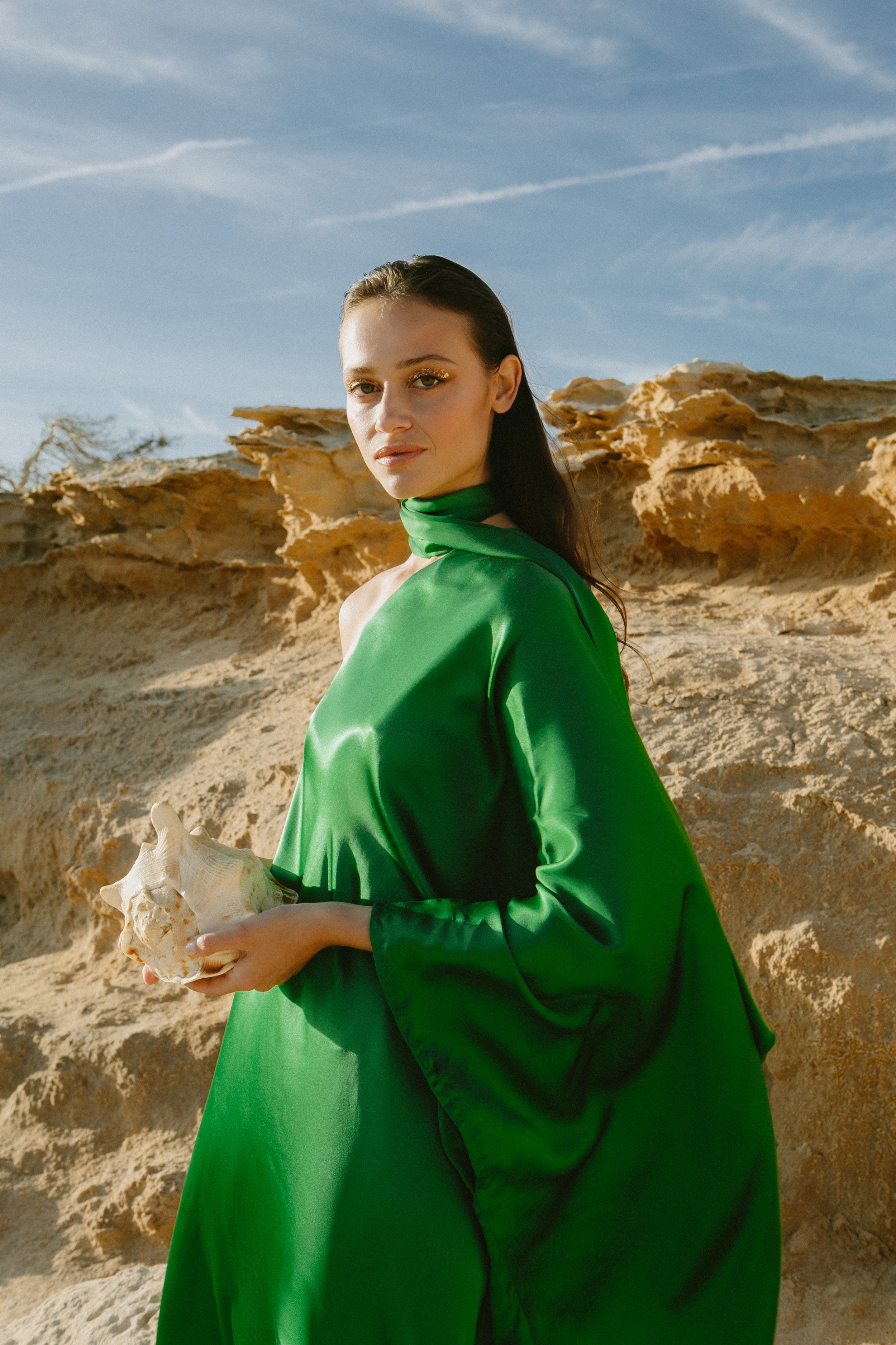 Woman in a green satin dress holding a seashell, standing against rocky desert landscape under a cloudy sky.