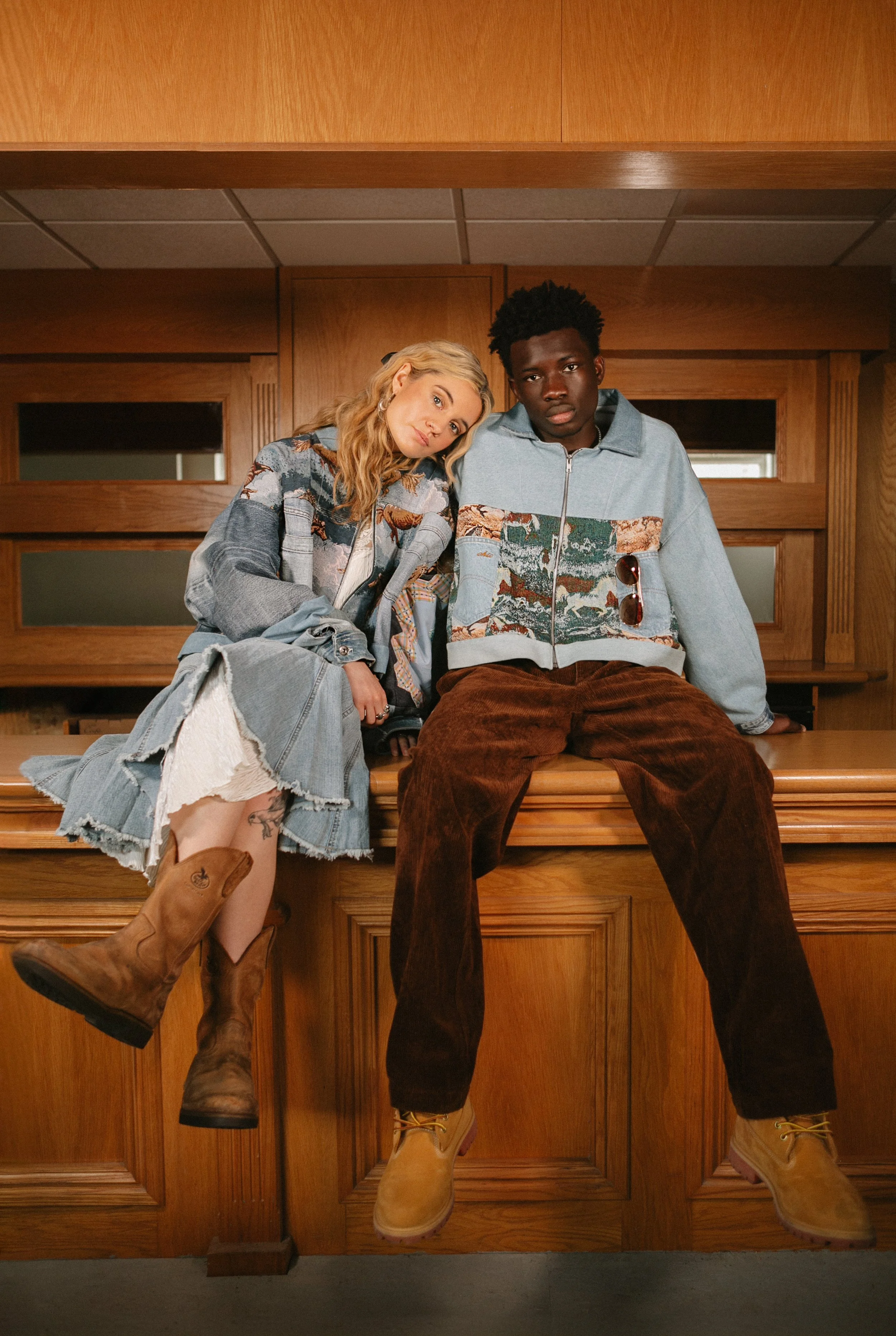 A young woman and man sitting on a wooden counter in a room with wooden paneling, dressed in casual jackets and boots, looking at the camera.