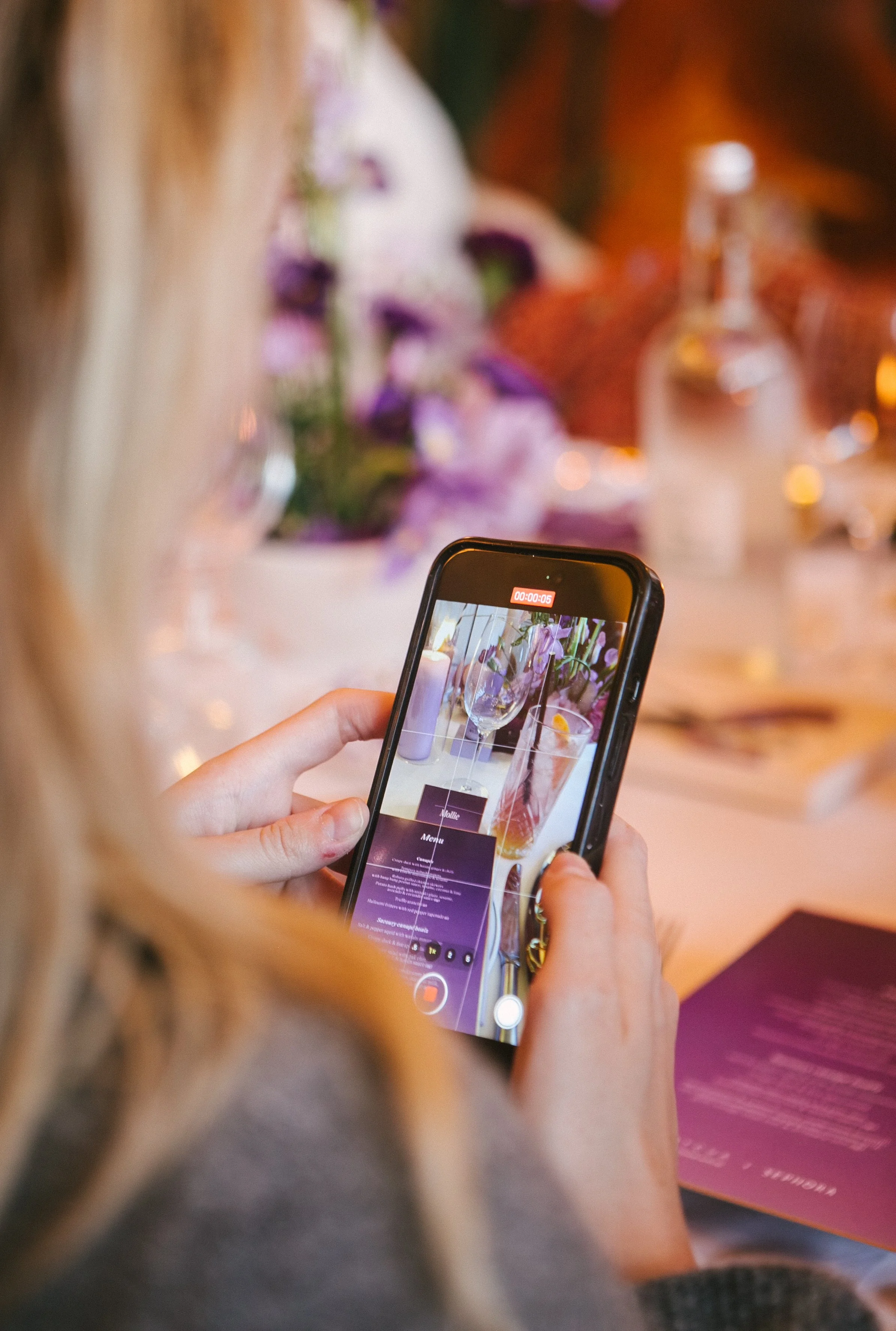 Person filming a decorative table setting with flowers and candles using a smartphone.