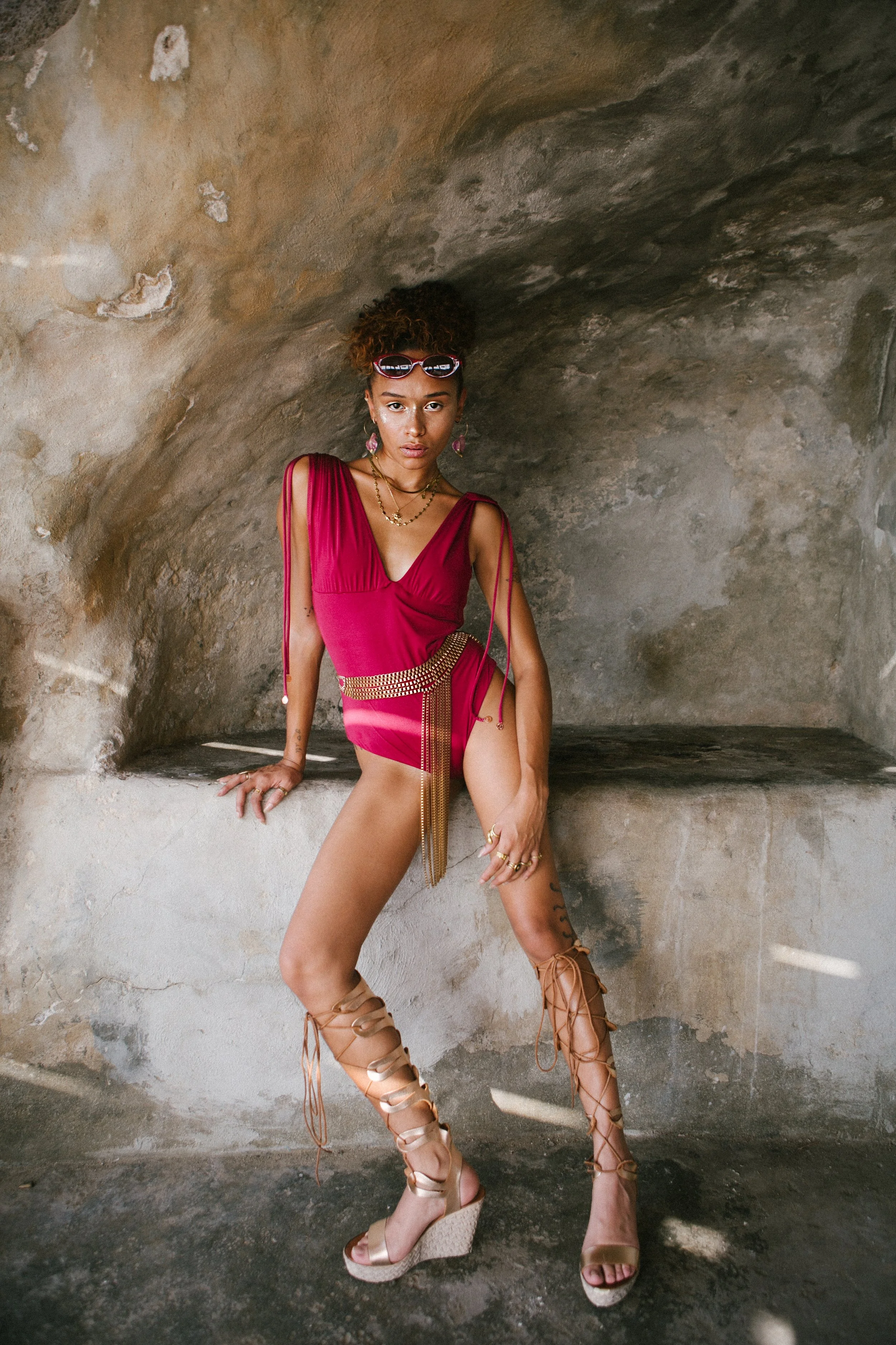 A woman in a vibrant red dress with gold accessories and tan lace-up wedge sandals posing inside a stone alcove.