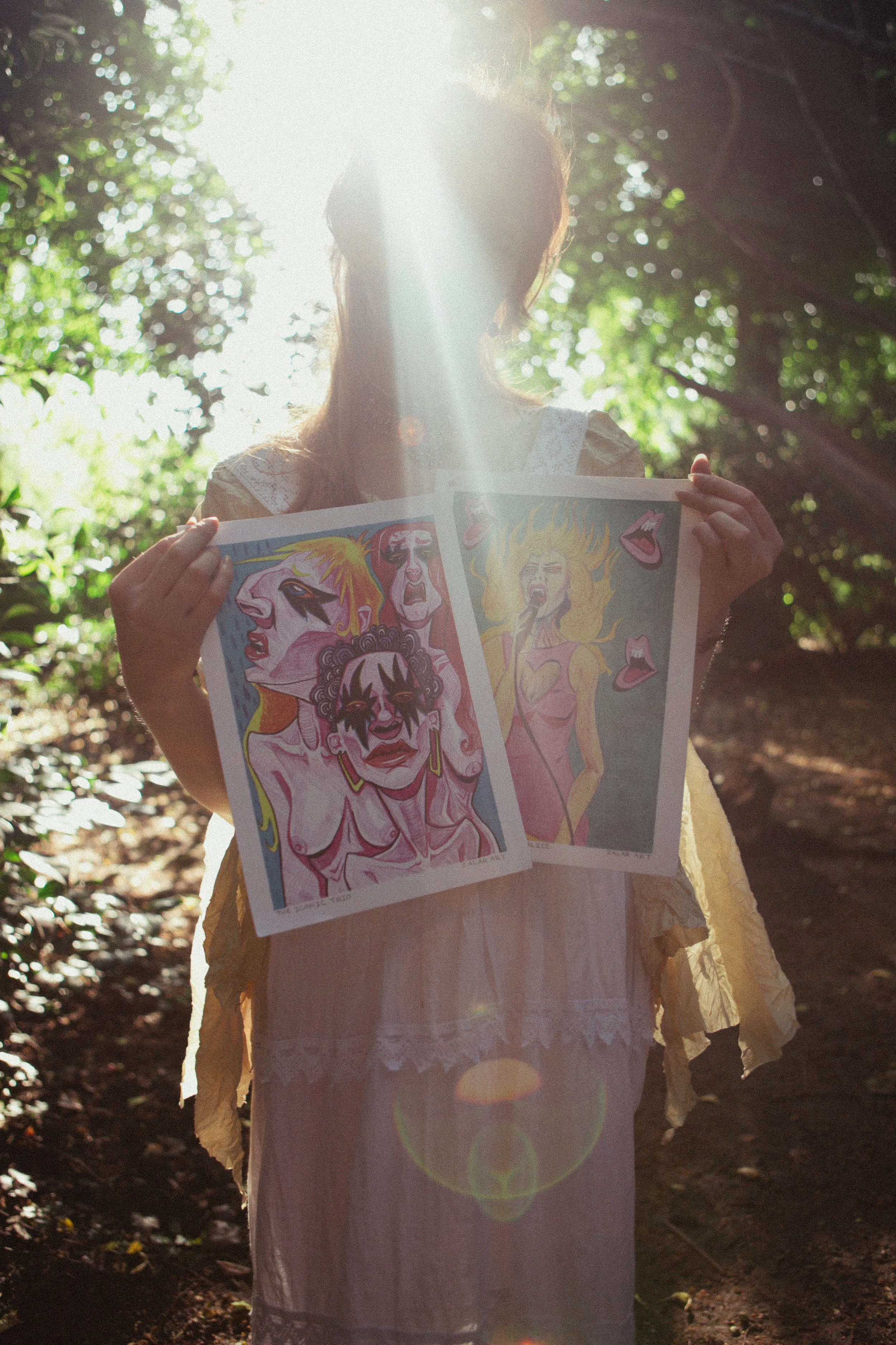 Person holding two colorful art prints outside in a wooded area with sunlight shining behind them.