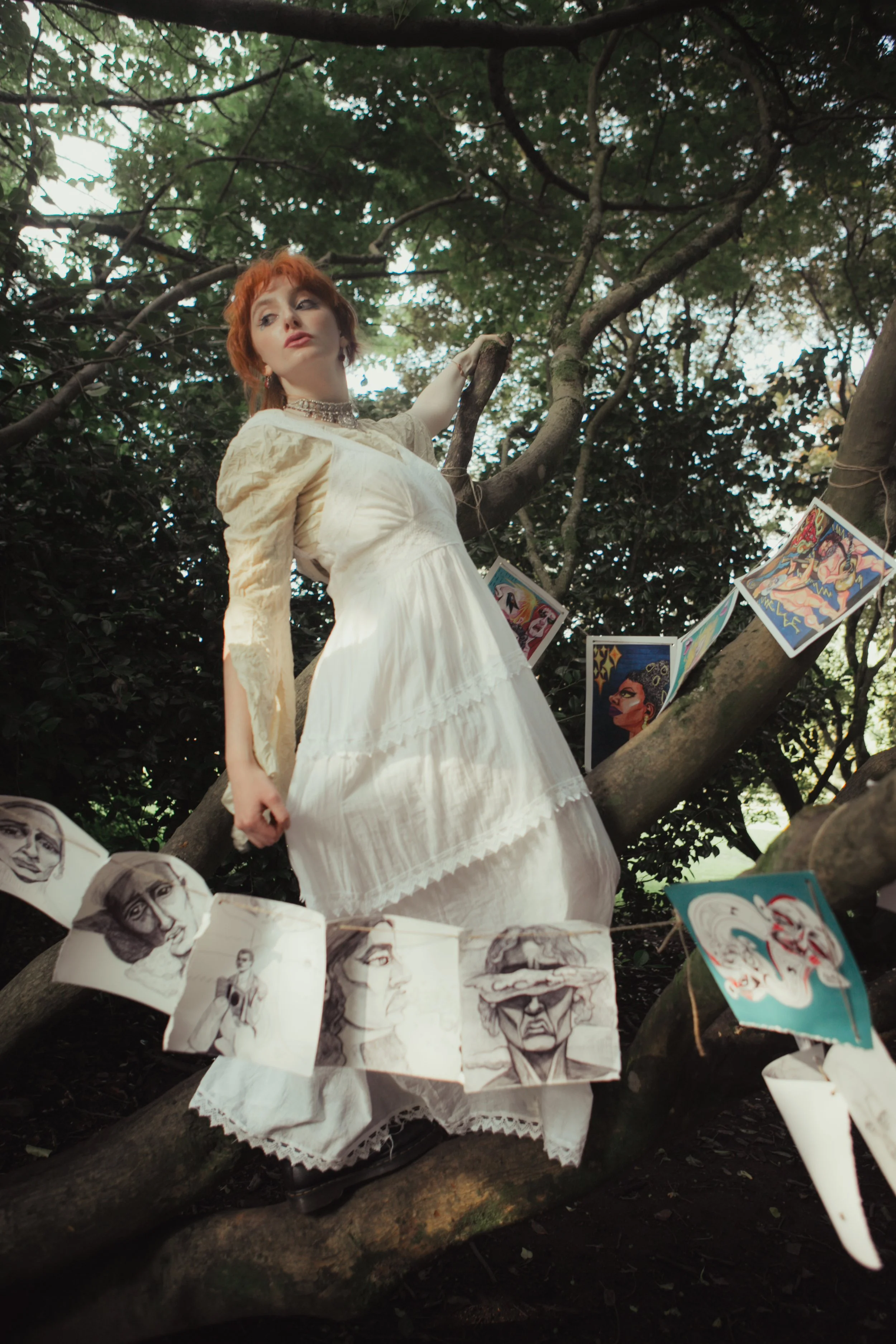 A woman with red hair wearing a white dress and jewelry, standing on a tree branch in a forested area, surrounded by hanging drawings and artwork.