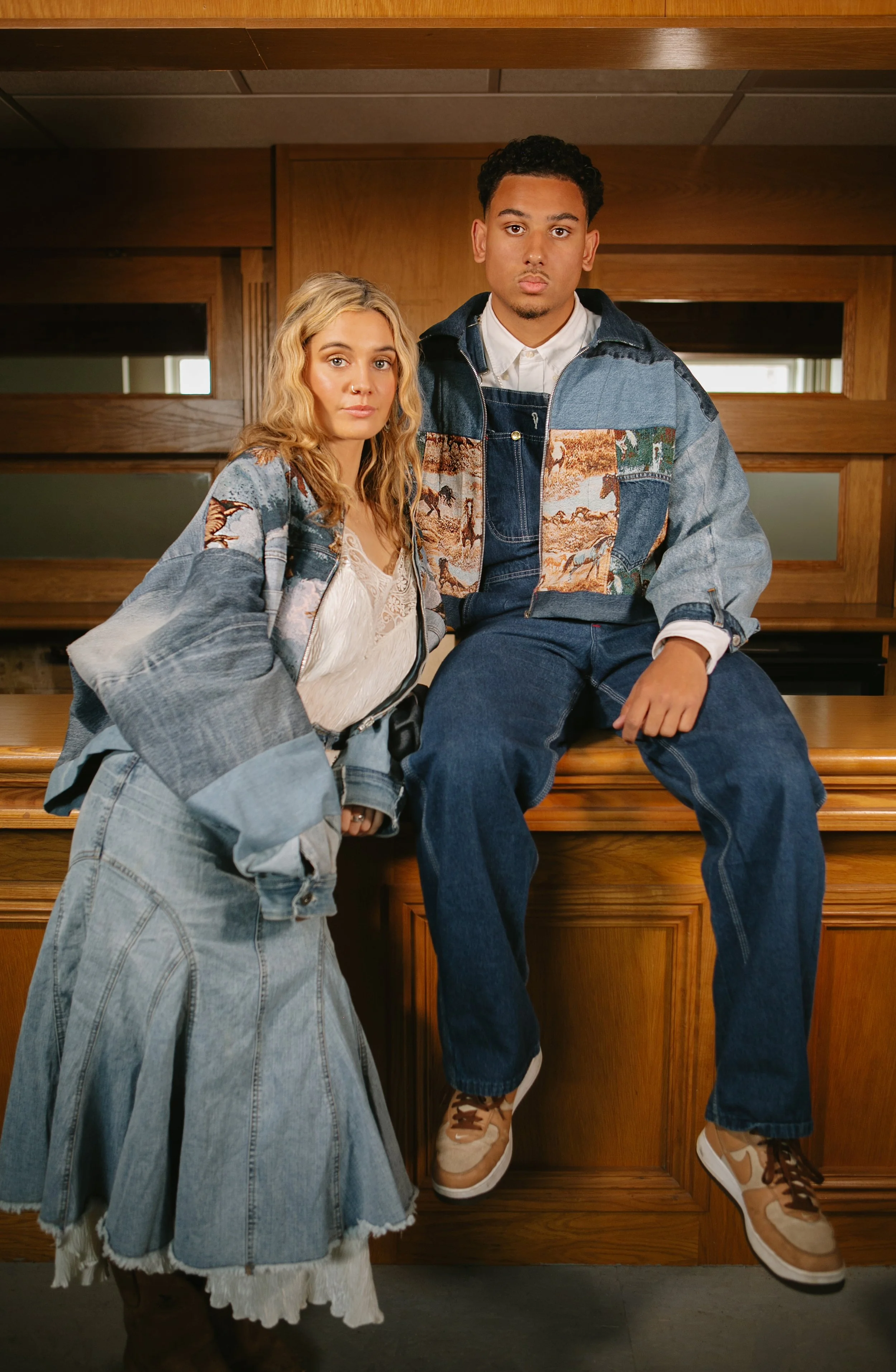 Two young adults, a woman and a man, sitting and standing on a wooden counter inside a room with wood paneling. They are dressed in layered denim clothing with artistic patches and prints. The woman has blonde curly hair and is wearing a large patchw