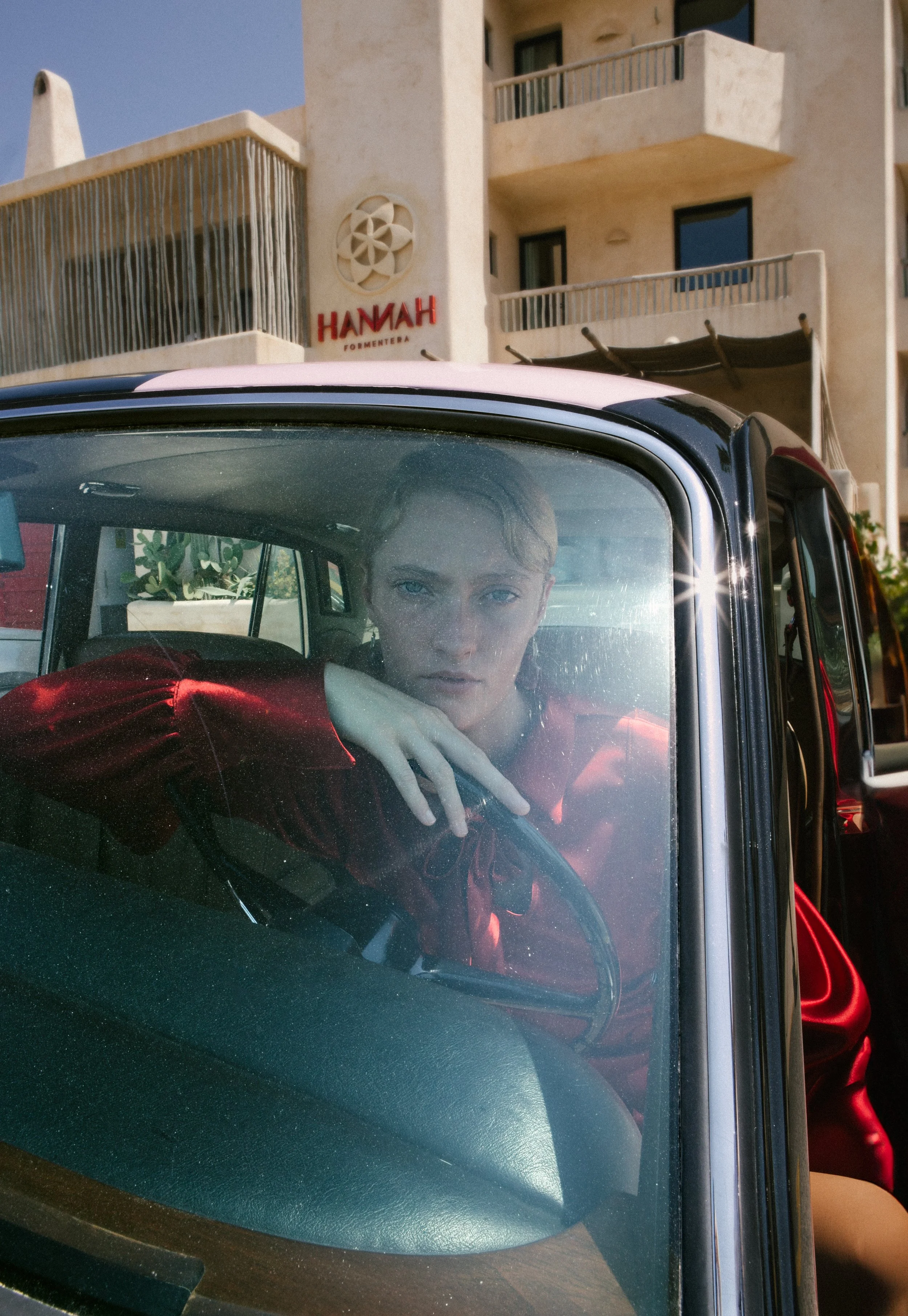 A person with short, blond hair and blue eyes sitting inside a vintage car, looking directly at the camera, with a building and hotel sign in the background.