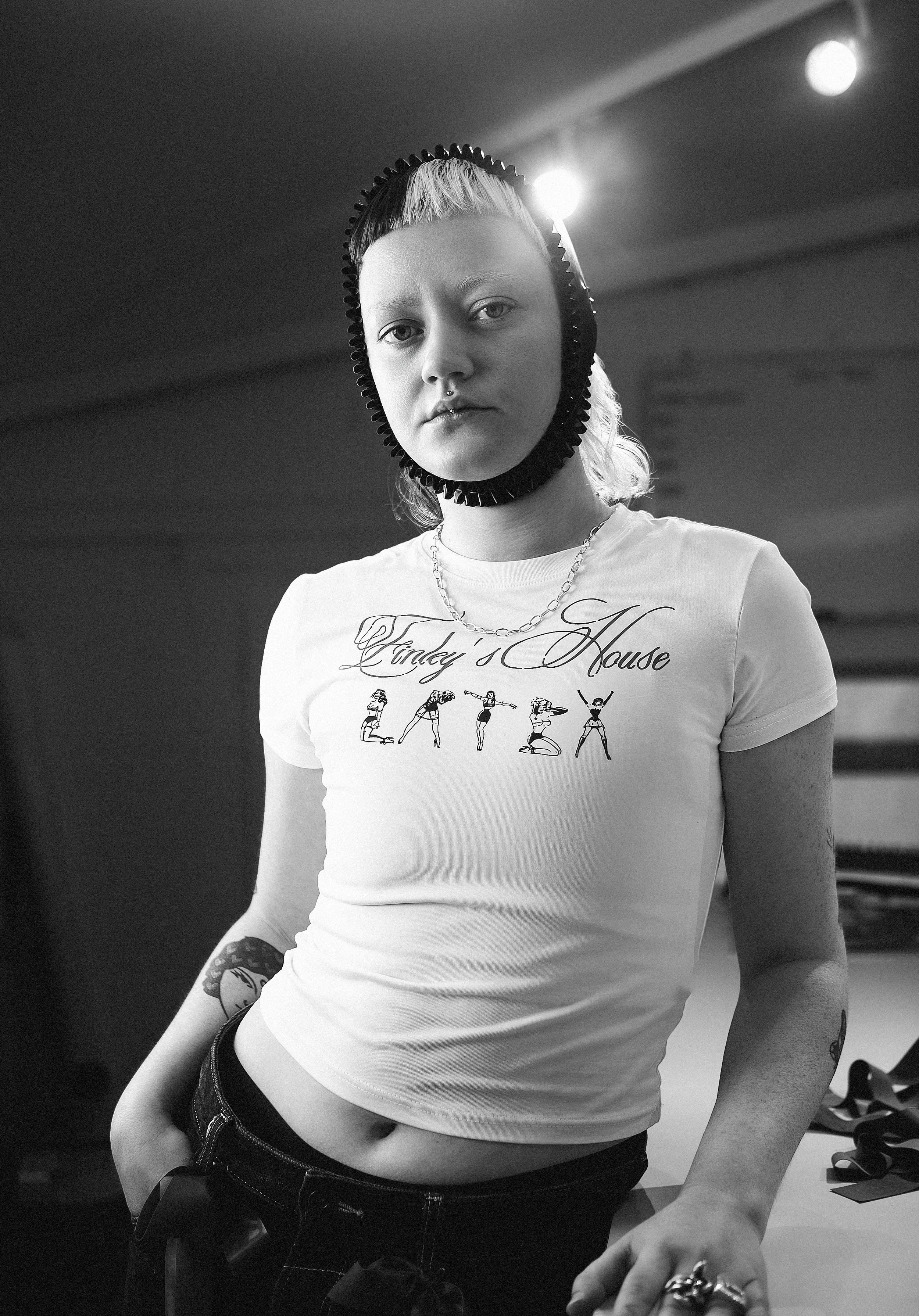 A young woman with a short blonde hairstyle wearing a head wrap with black trim. She is dressed in a white T-shirt with the text 'Cindy's House' and illustrations of yoga poses, paired with dark jeans. She has tattoos on both arms and wears rings, a necklace, and has a serious expression, standing in an indoor setting with visible light fixtures.