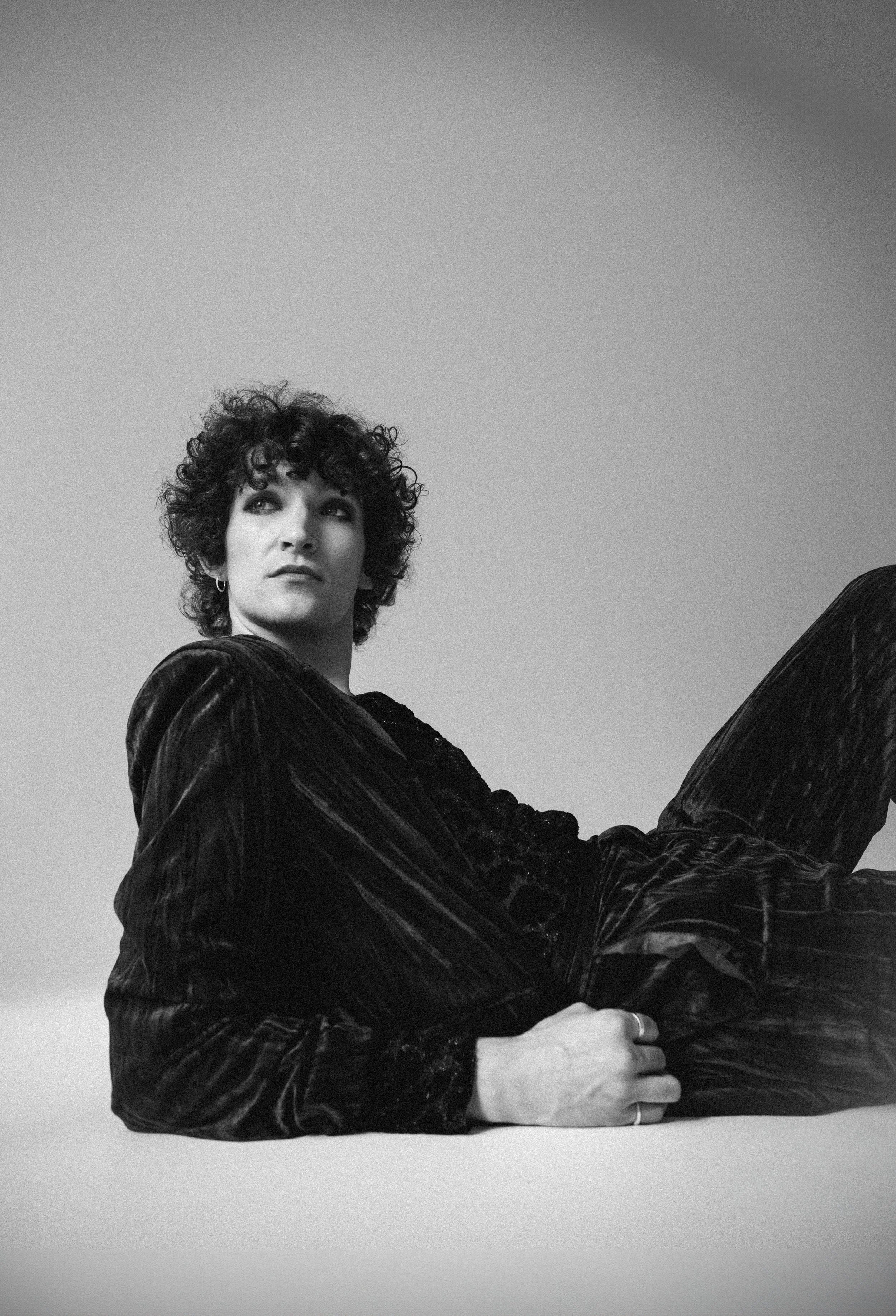 Black and white photo of a person with curly hair, wearing a dark velvet outfit, reclining against a plain background, looking to the side.
