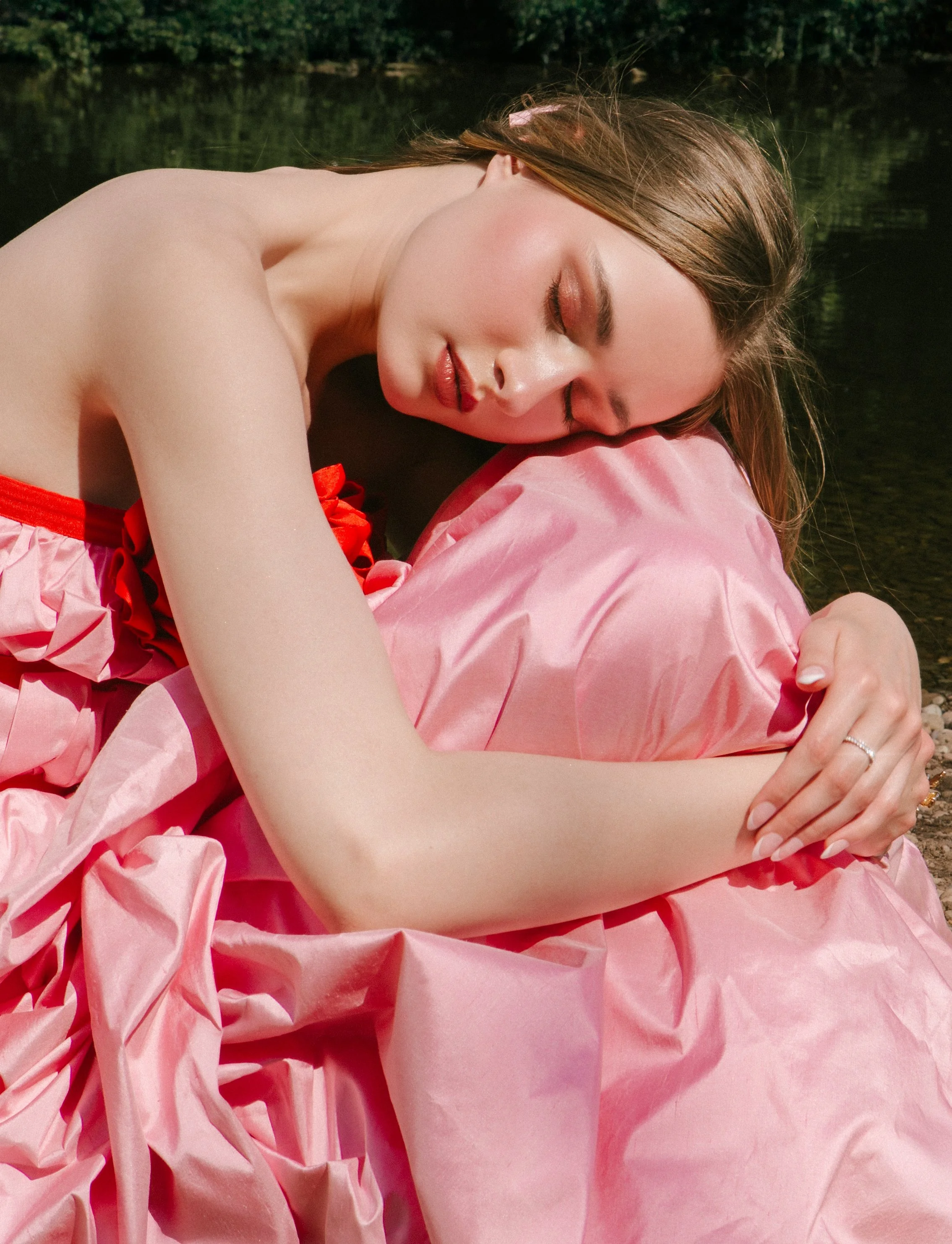 A woman with long brown hair and makeup, wearing a pink dress with ruffles, is lying on a pink silky fabric with her eyes closed, resting her head on her arm by a body of water with greenery in the background.