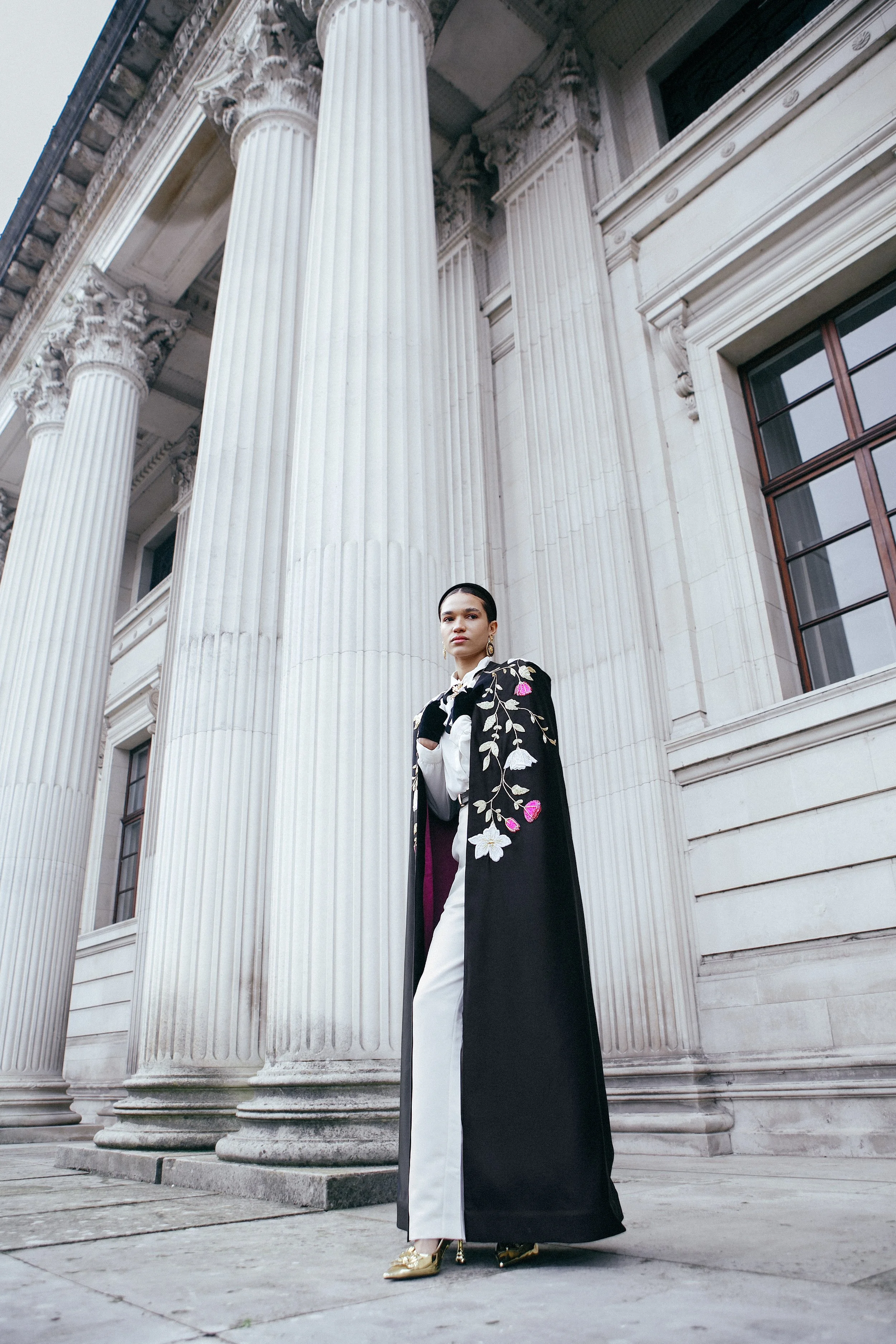 A woman wearing a long black embroidered coat over white pants and gold shoes standing in front of a grand classical building with large columns.