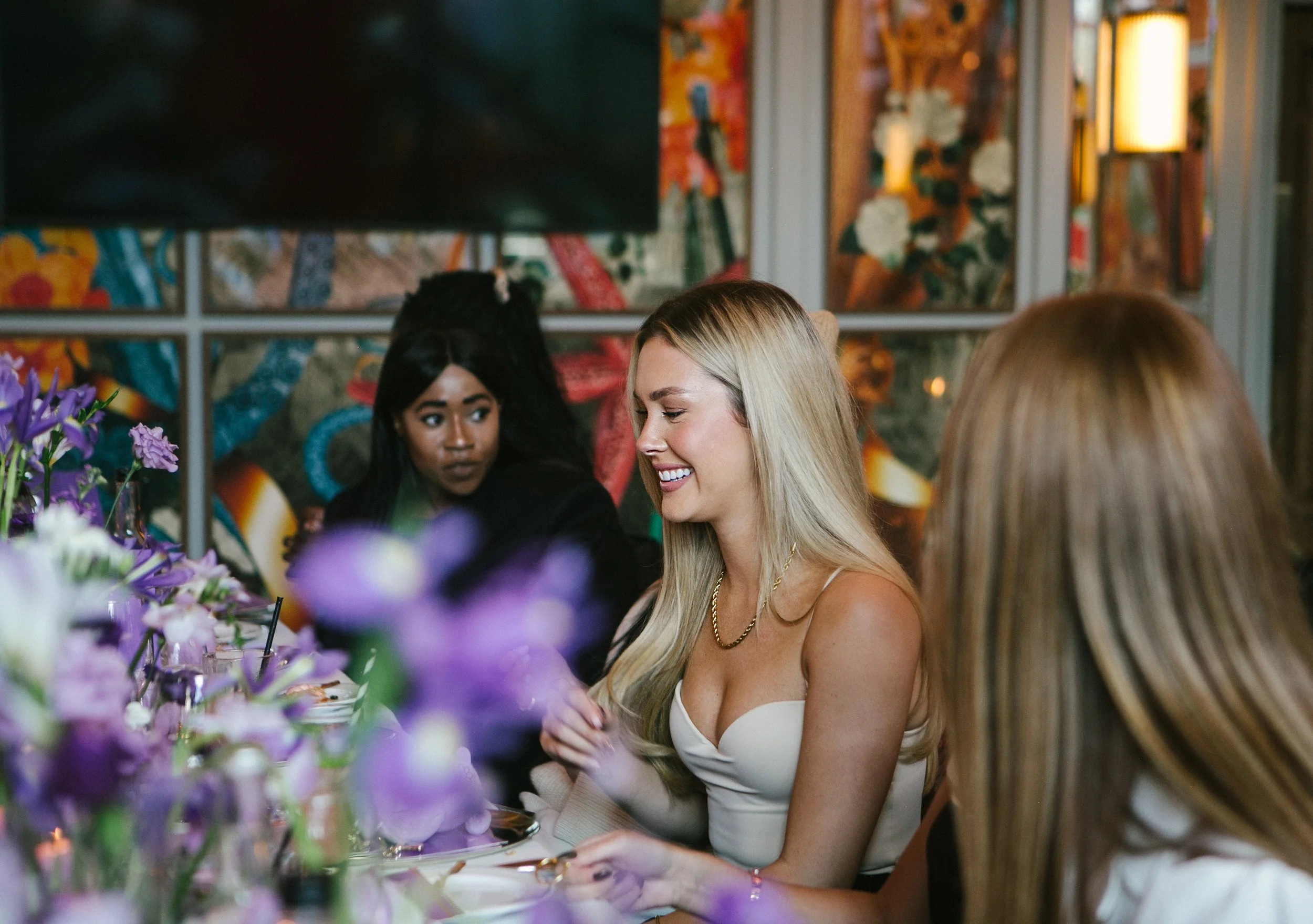 A group of women sitting at a table decorated with purple flowers in a lively restaurant or cafe with colorful artwork on the walls, engaged in conversation with one woman smiling.