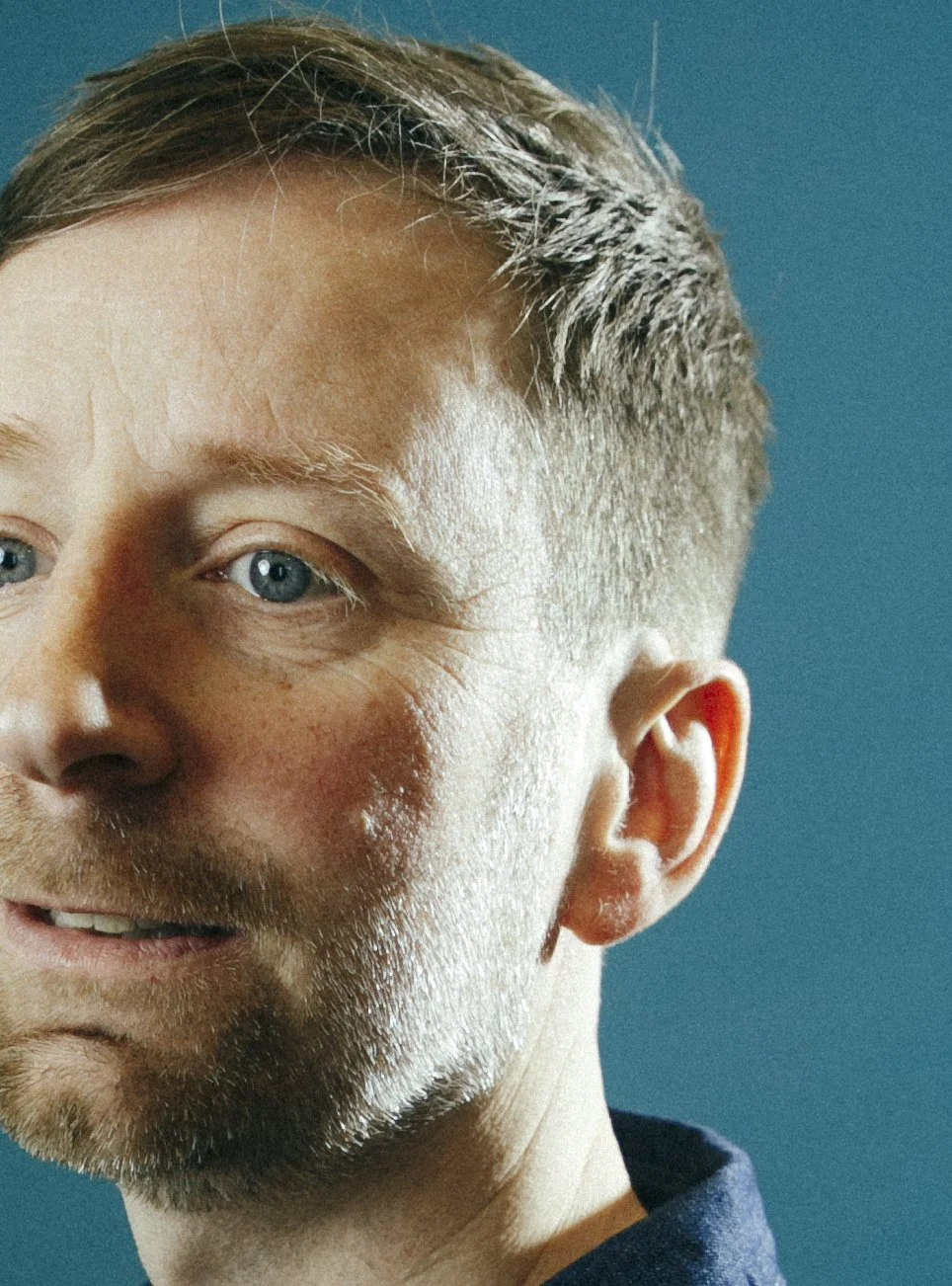 Close-up of a man's face showing his left eye, partial nose, ear, and smiling mouth against a blue background.