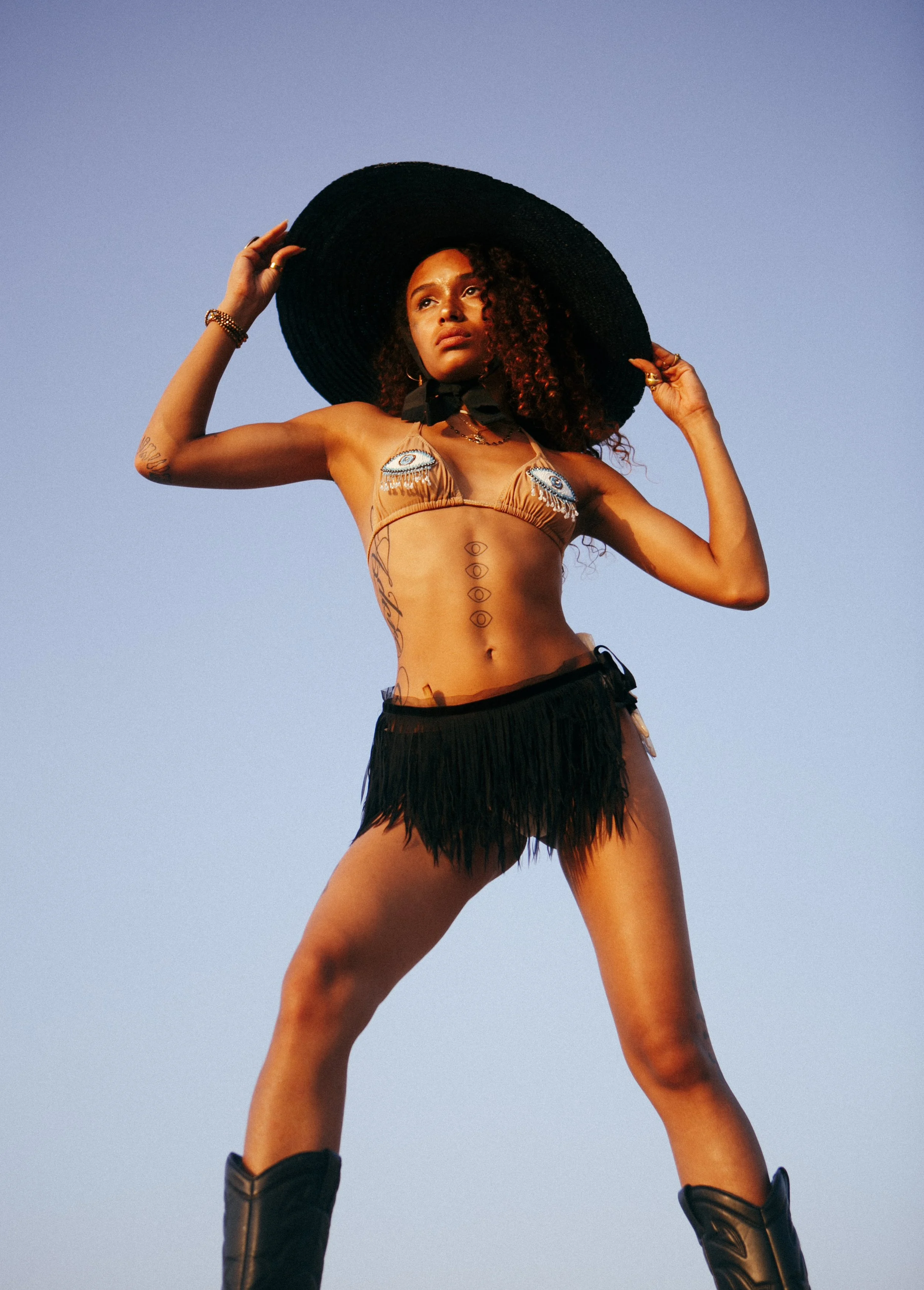 A woman wearing a large black sunhat, a bikini top with eye designs, a black fringed skirt, and black boots, standing outdoors against a clear blue sky.