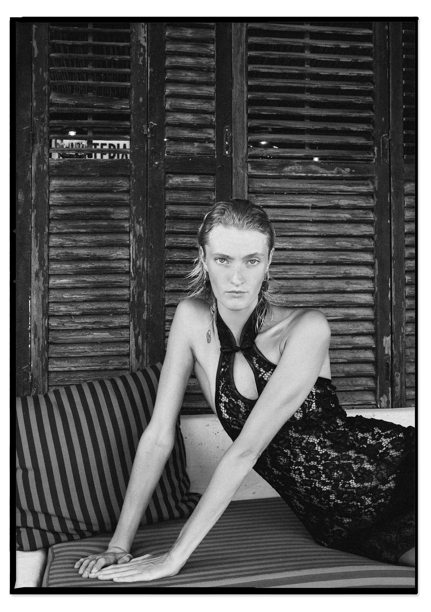A woman in a black lace dress posed on a striped bench with wooden shutters in the background.
