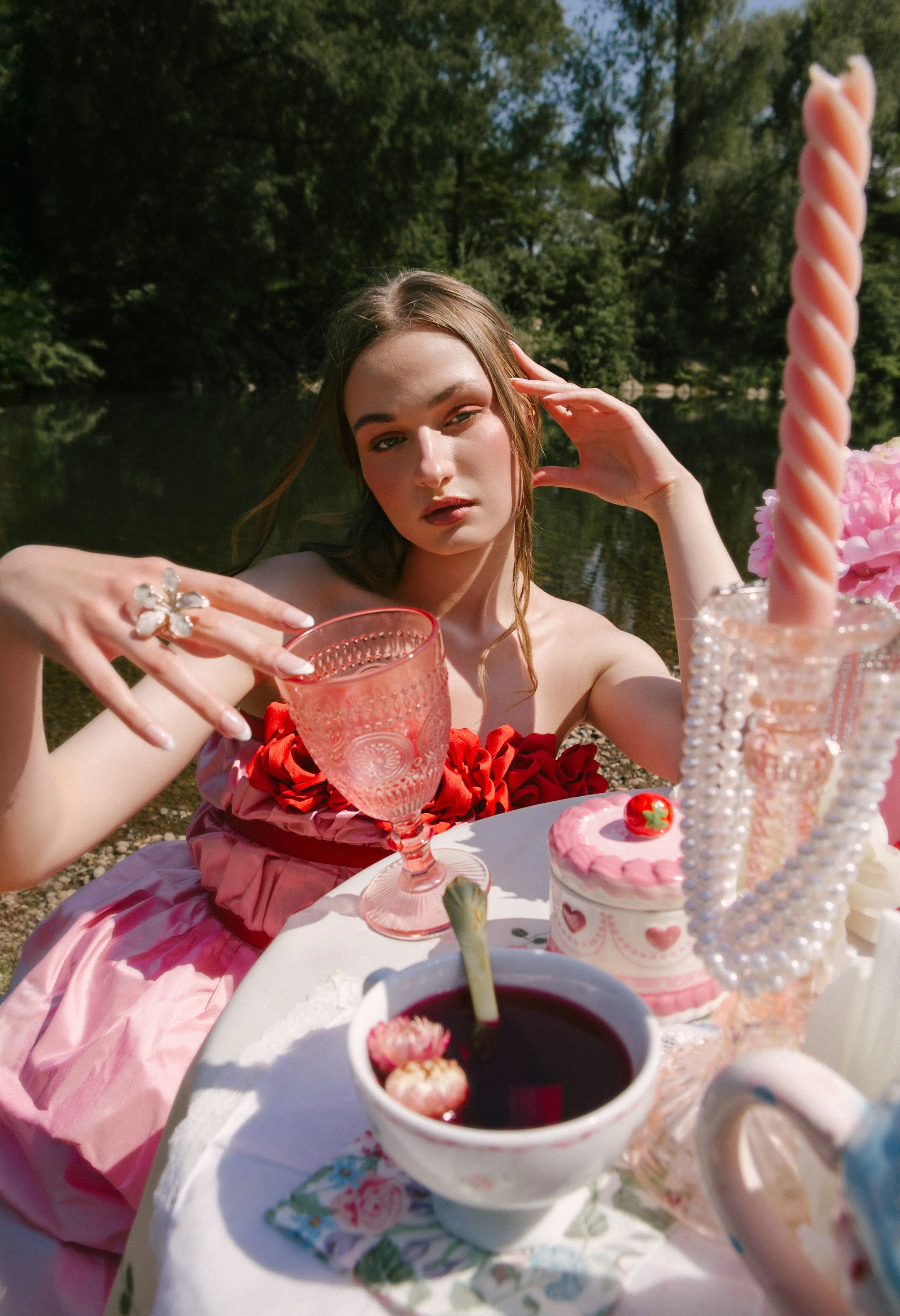 A young woman in a pink dress sitting at a table outdoors near a body of water with trees in the background, surrounded by tea cups, a cake, a teapot, and decorative items.