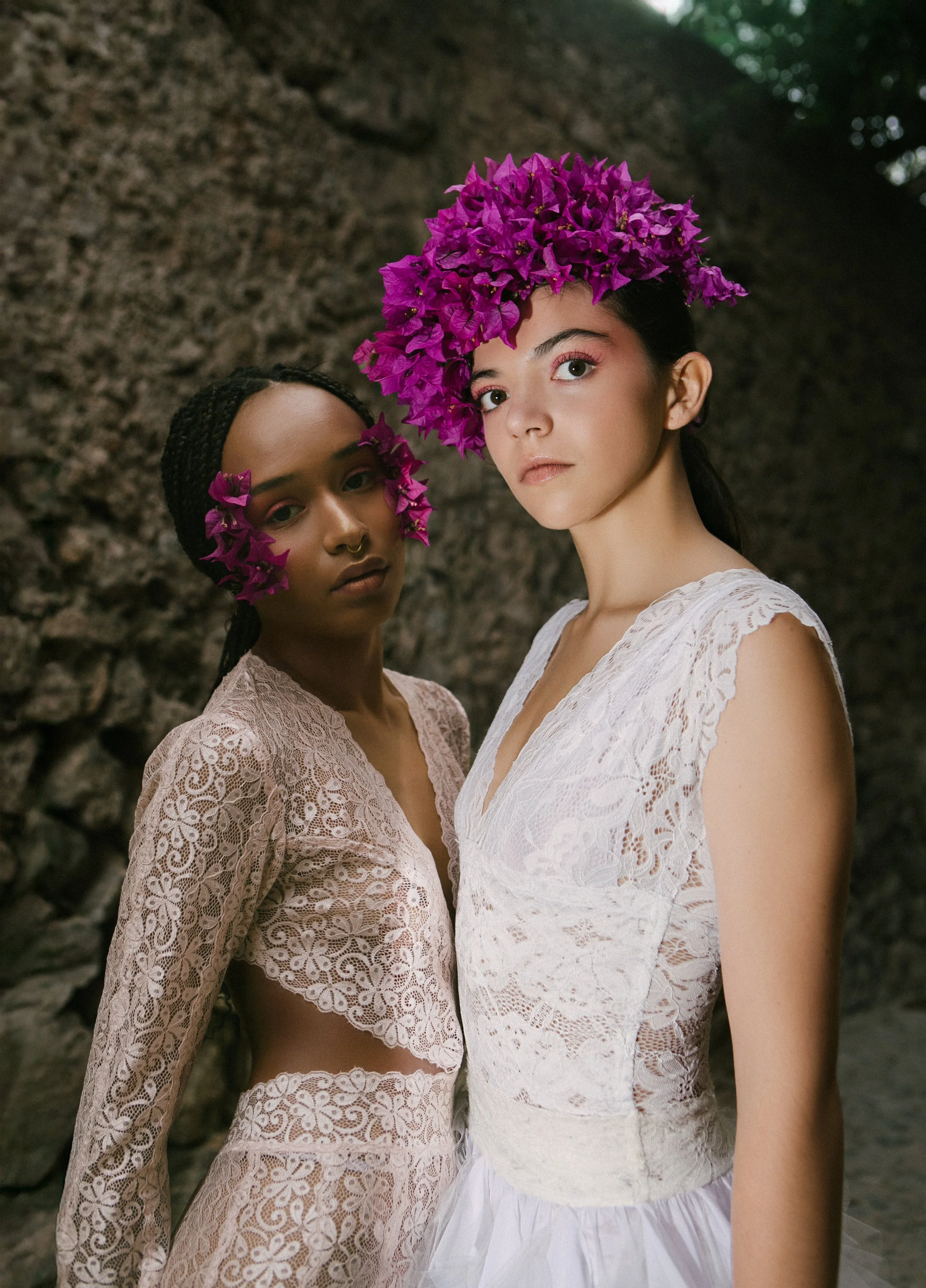 Two women with flower headpieces and lace dresses standing outdoors in front of a rocky background.