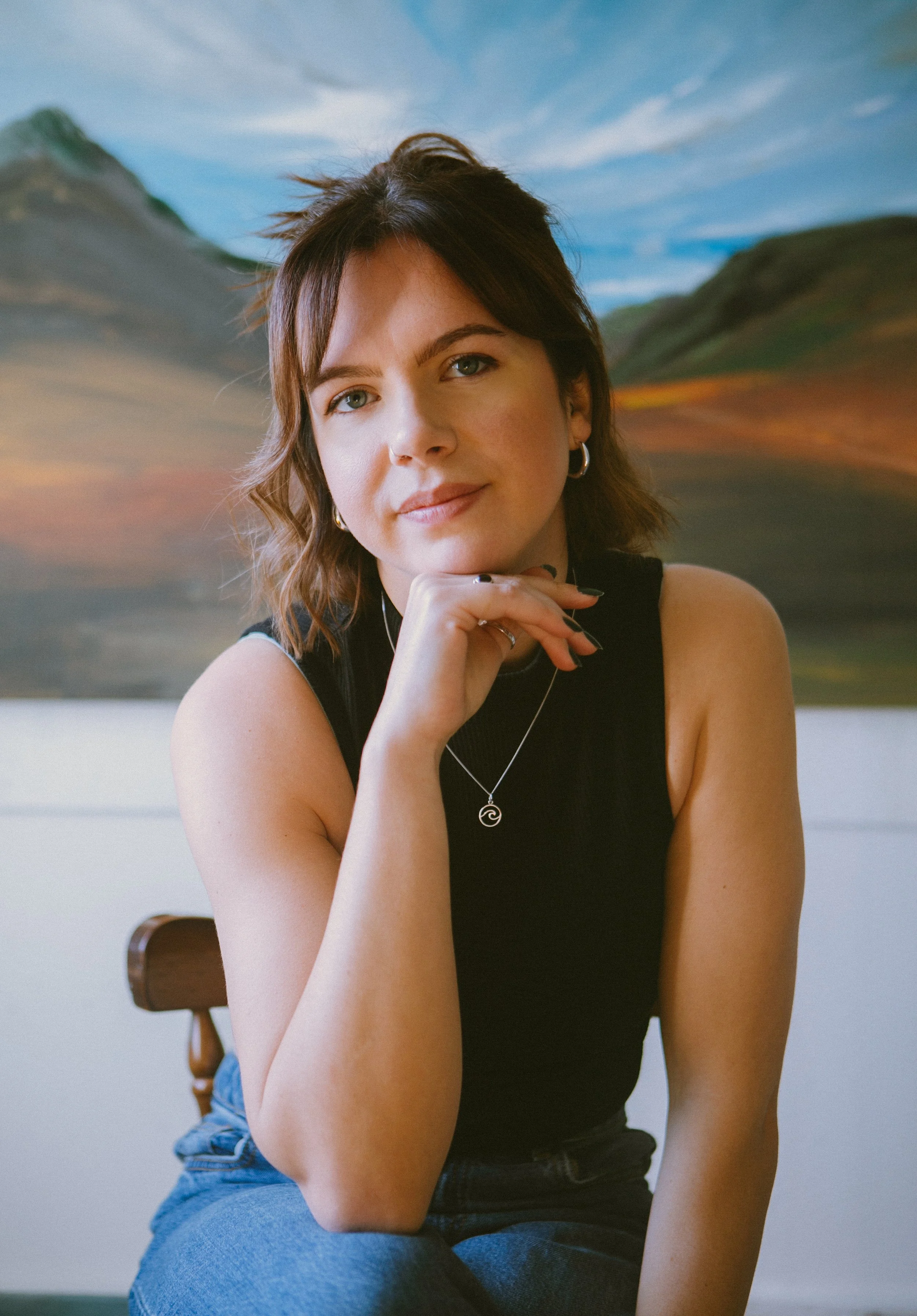 A young woman with shoulder-length brown hair and earrings sits in front of a landscape painting. She is wearing a black sleeveless top and layered necklaces, with her chin resting on her hand, looking at the camera.