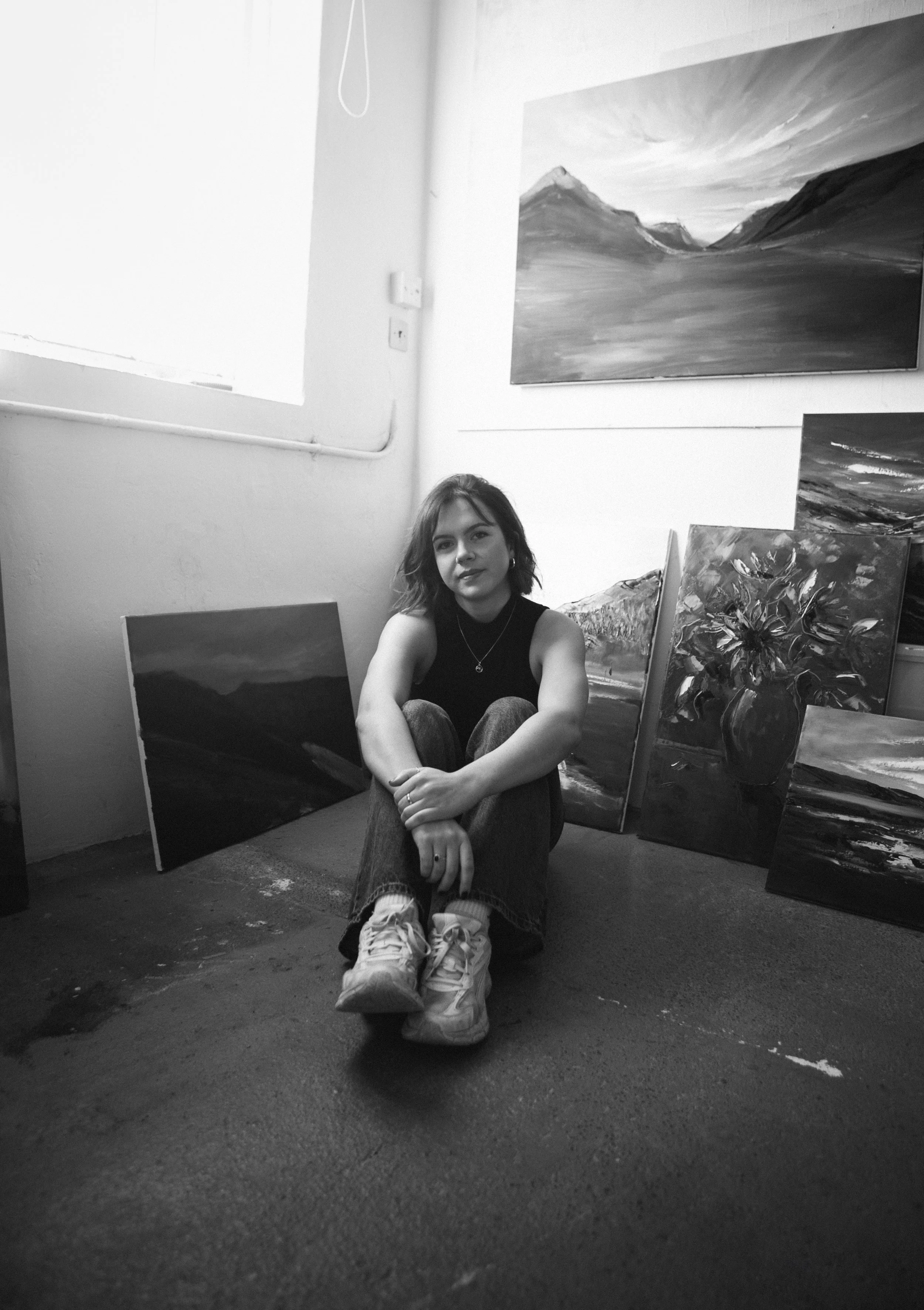 A young woman sitting on a floor surrounded by landscape paintings in an art studio.