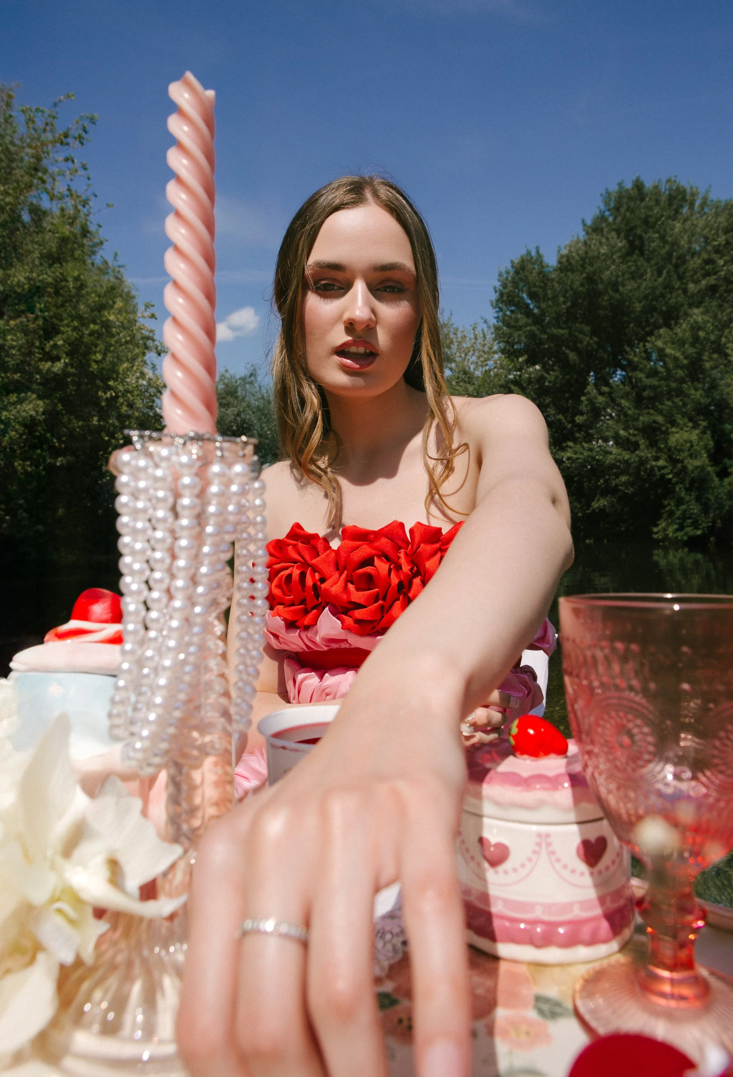 Young woman with long hair wearing a red floral dress, sitting at an outdoor table with desserts and decorations, holding her hand towards the camera, with a blue sky and green trees in the background.