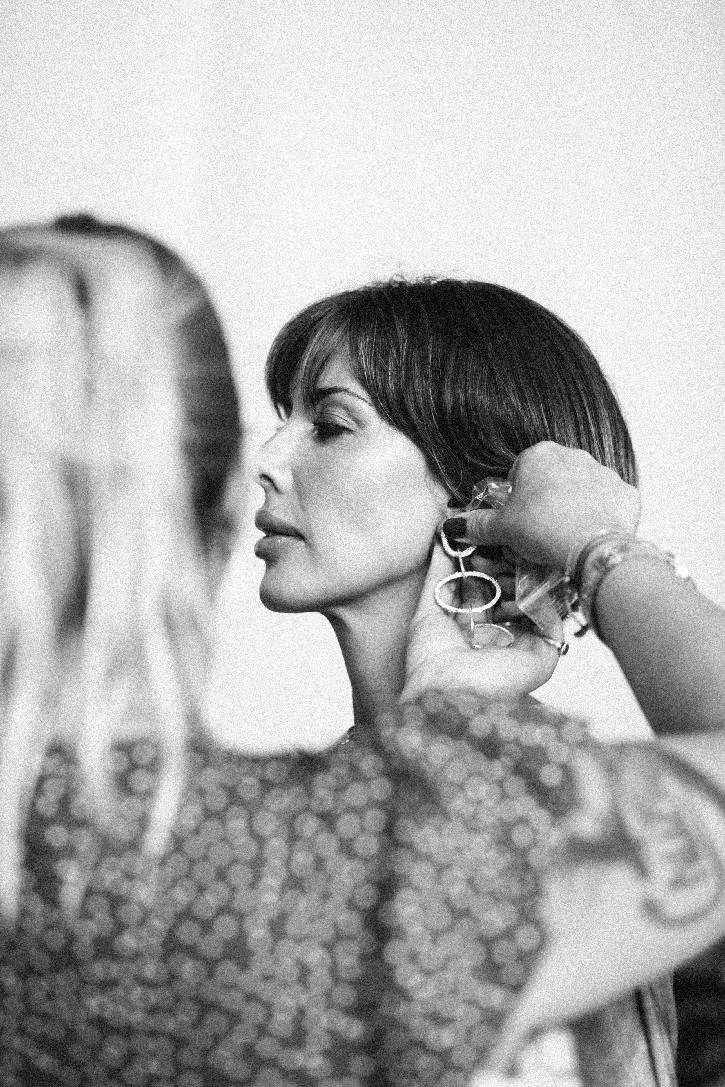A woman with short dark hair getting her ear pierced by a professional, while another person, with long blonde hair, is partially visible in the foreground.
