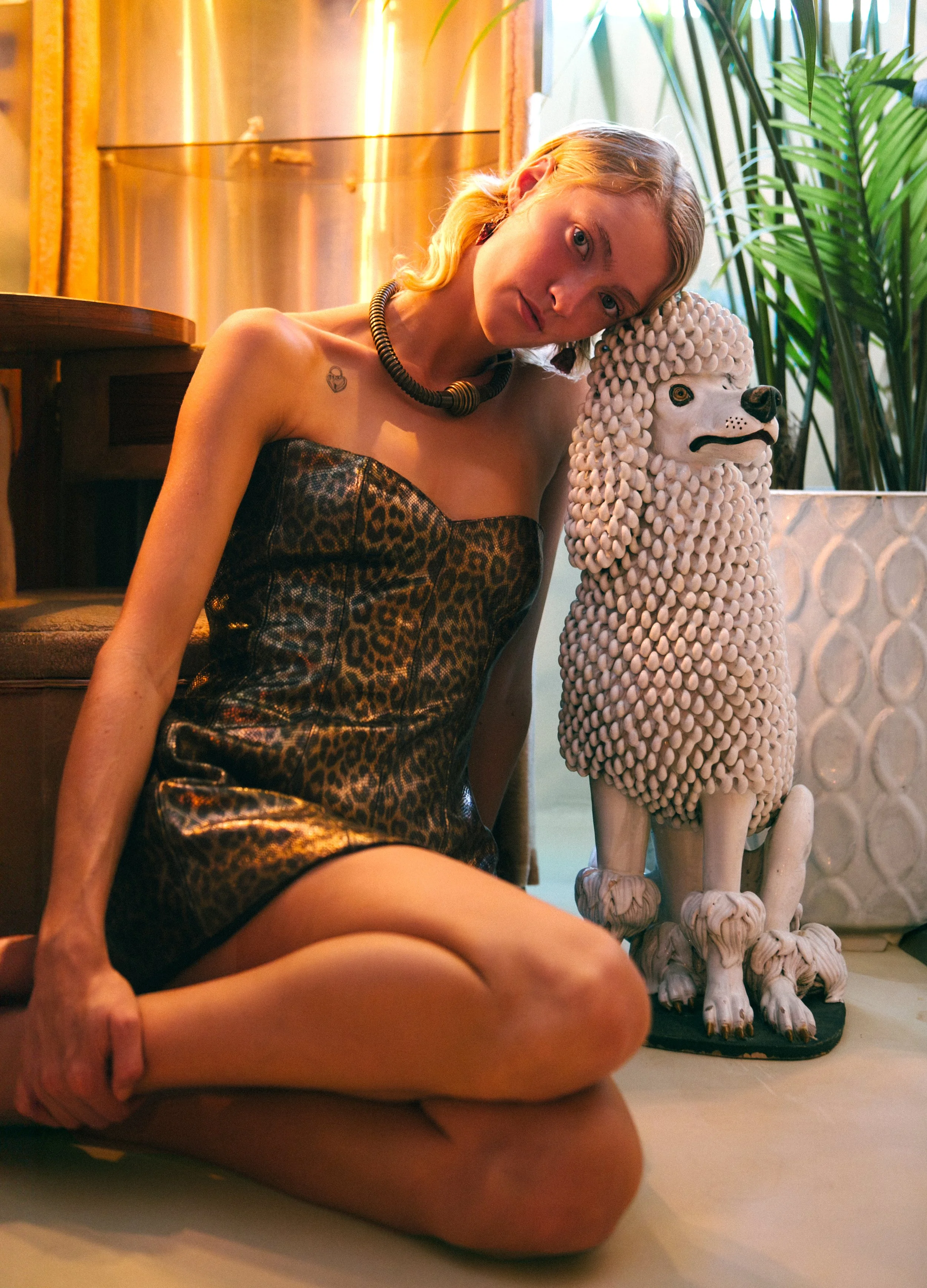 Young woman with blonde hair wearing a leopard-print strapless dress and a black necklace, sitting on the floor with her knees up and leaning her head on a ceramic statue of a poodle dog with a curly textured coat, in a room with plants and warm ligh