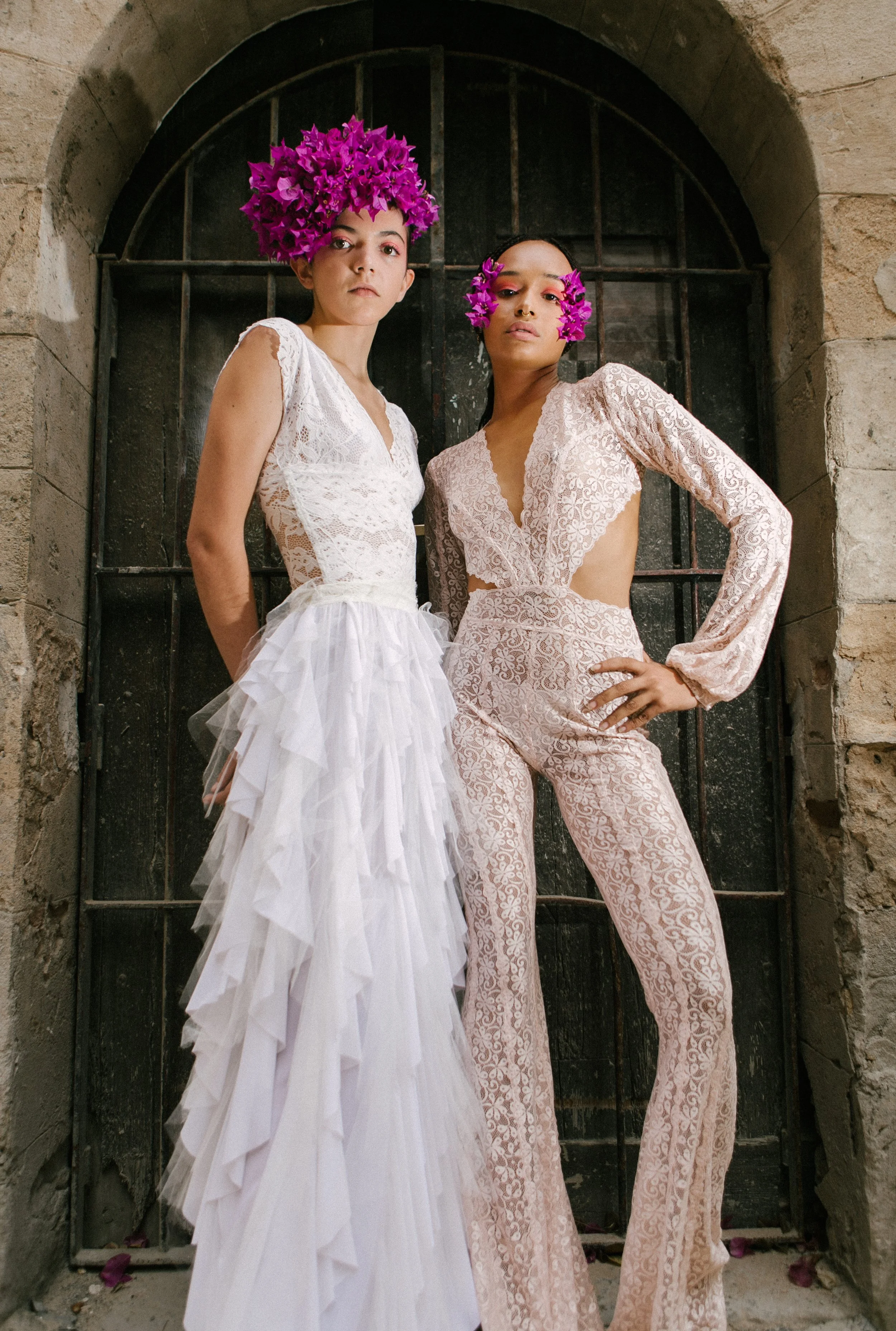 Two women posing in front of a black iron gate, wearing lace dresses and purple flower headpieces; one in a white wedding dress and the other in a pink lace jumpsuit.
