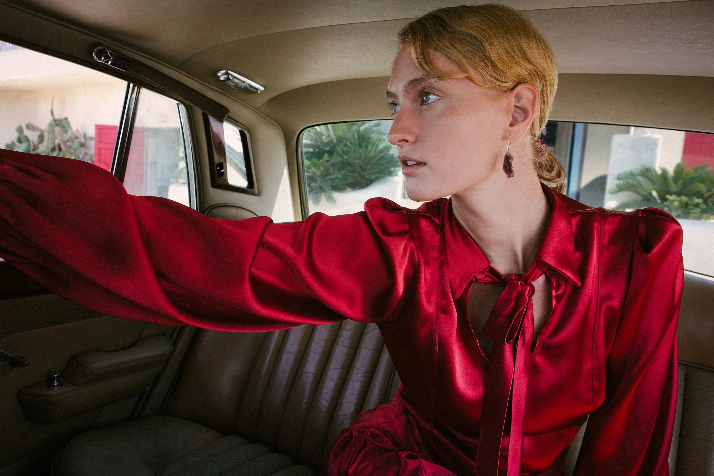 A woman with blonde hair wearing a shiny red blouse and earrings sitting in the backseat of a vintage car, looking out the window.
