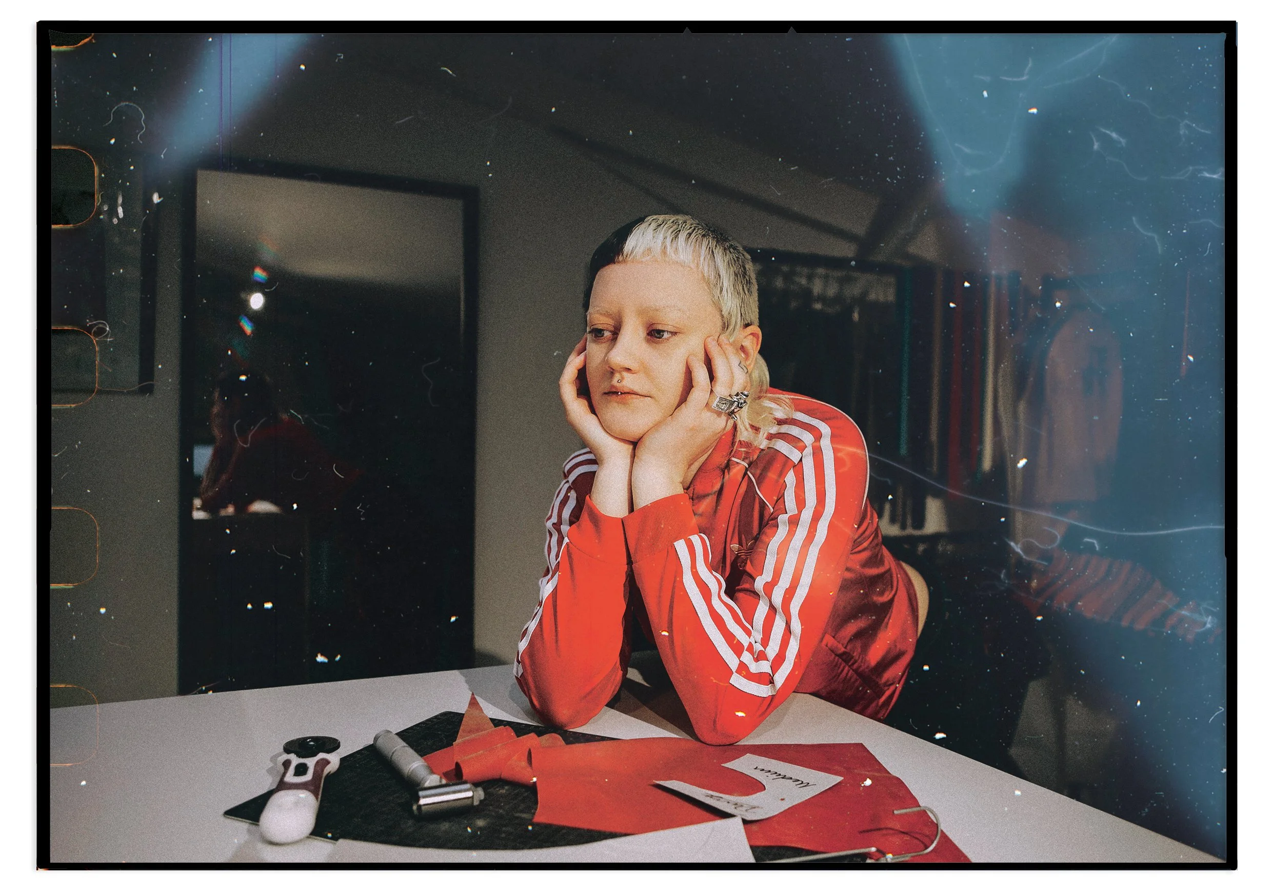 Person with short, two-toned hair (blonde and black) resting their face on hands at a table, wearing a red Adidas jacket with white stripes, with some art supplies and papers on the table.