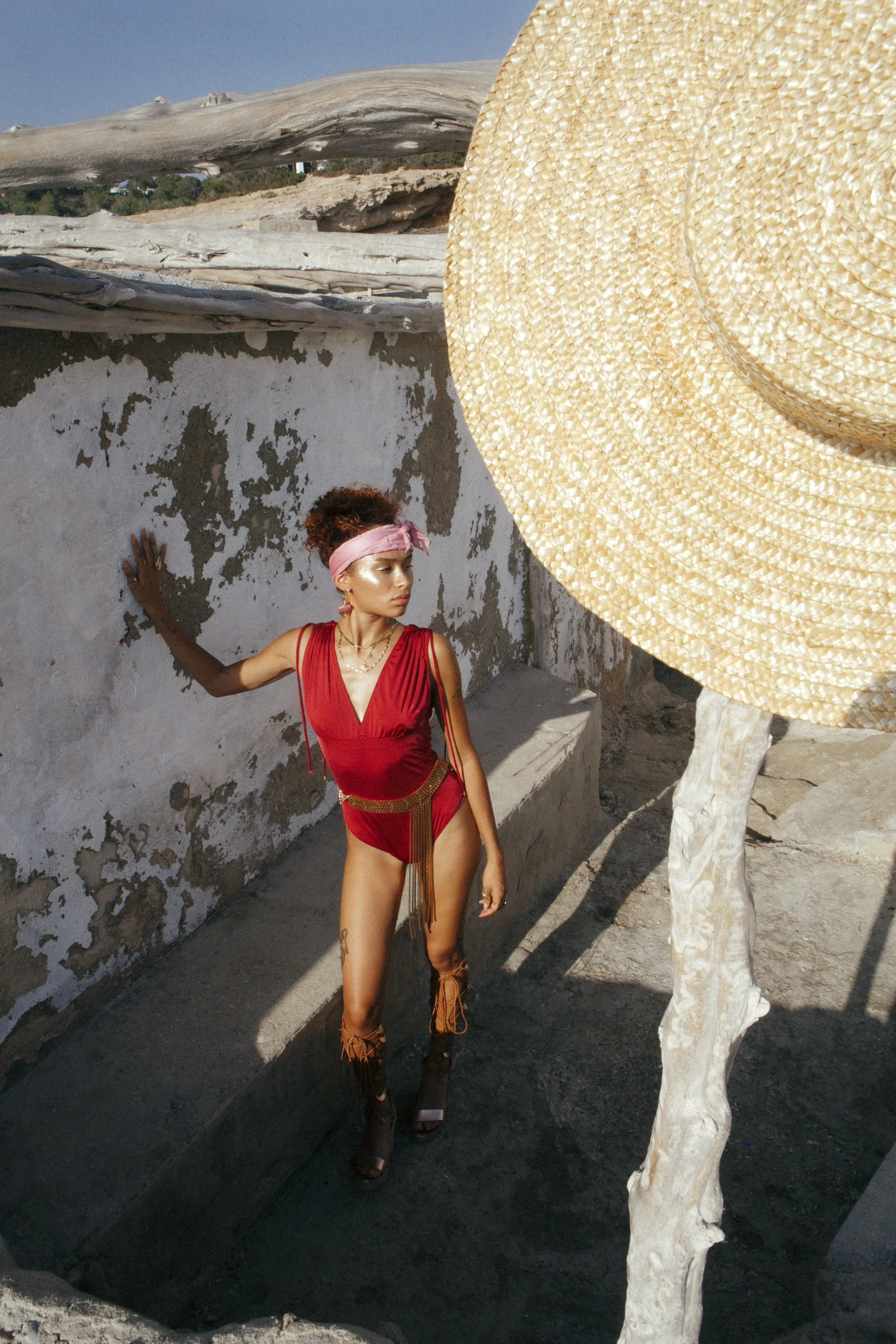 A woman in a red outfit and pink headband leaning against a weathered wall, with a large straw hat in the foreground.