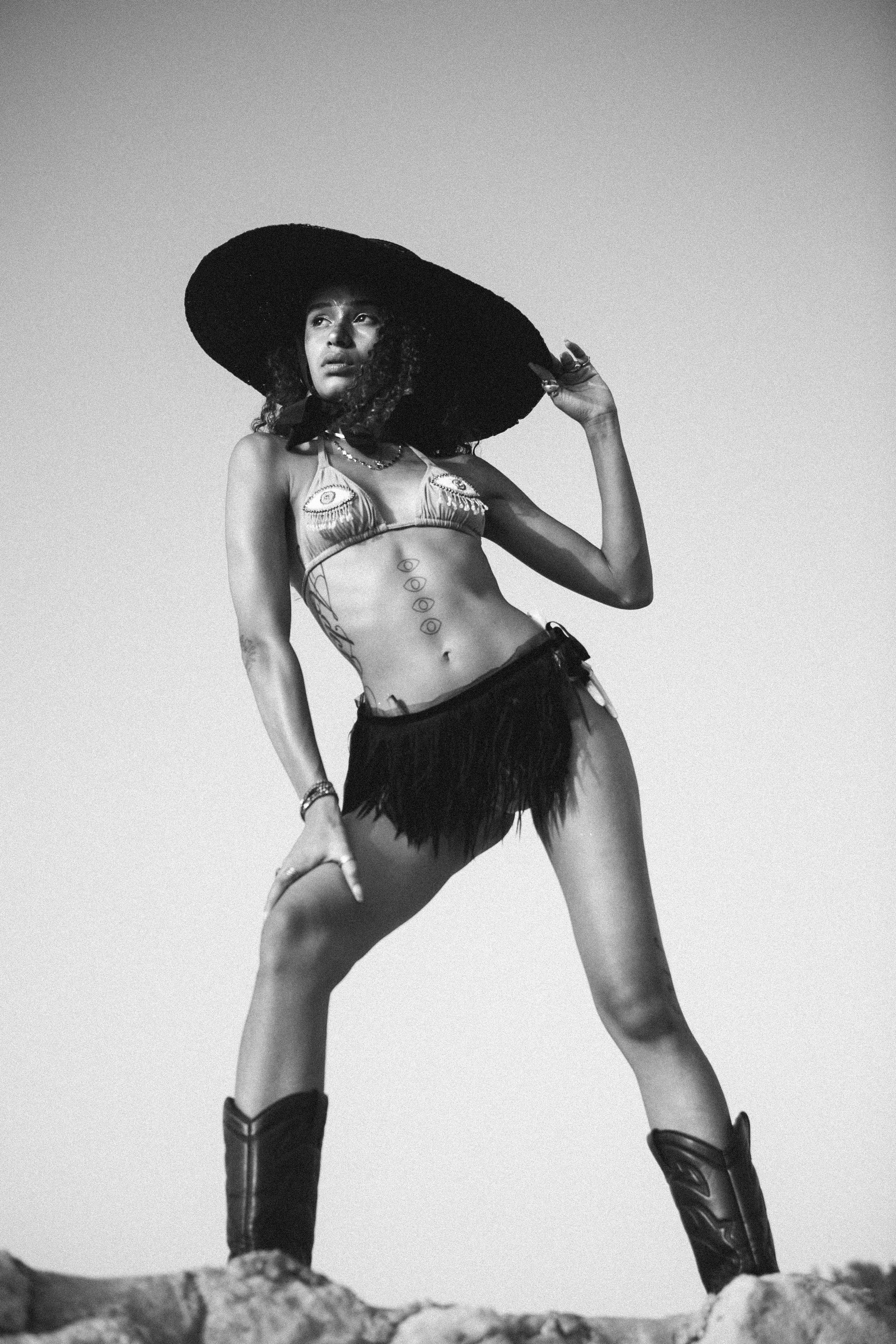 A woman standing outdoors wearing a large sun hat, a bikini top with eye designs, fringe shorts, and cowboy boots, striking a confident pose.