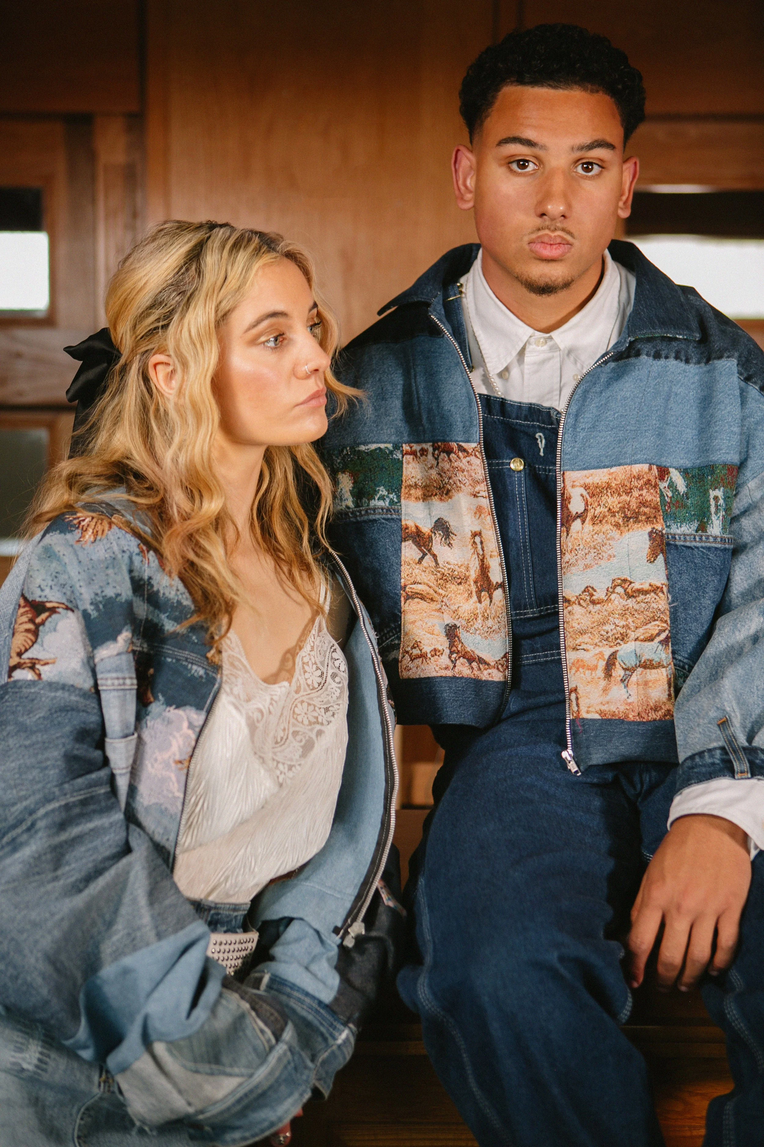 Two young adults, a woman with curly blonde hair and a man with short curly hair, sitting indoors with serious expressions. Both are wearing denim jackets with artistic prints. The woman is in a white lace top with a denim jacket, and the man is in a