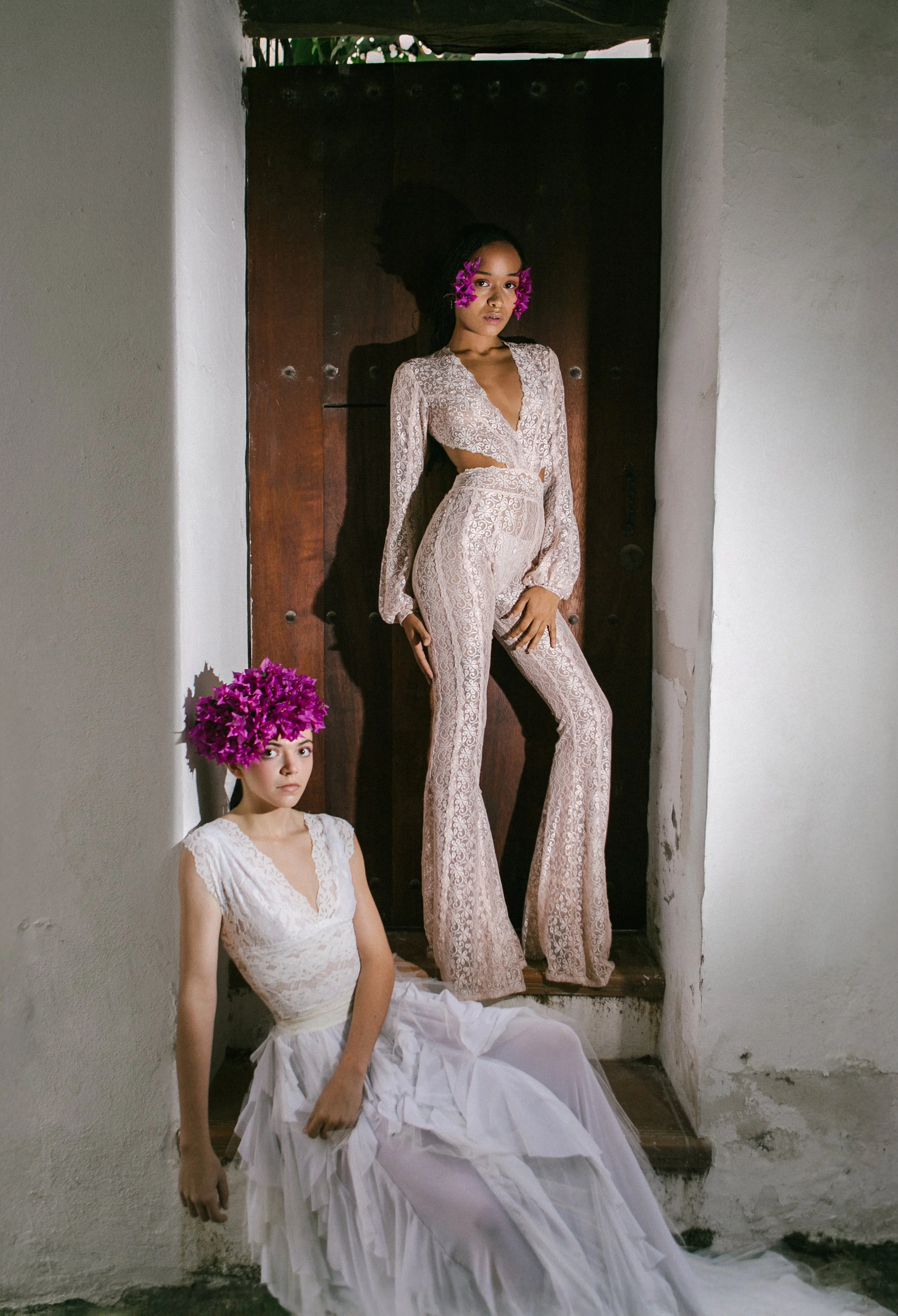 Two women wearing white lace clothing and purple floral headpieces, one standing on a ledge and the other sitting on the floor, in a minimalist indoor setting with white walls and a dark wooden door.