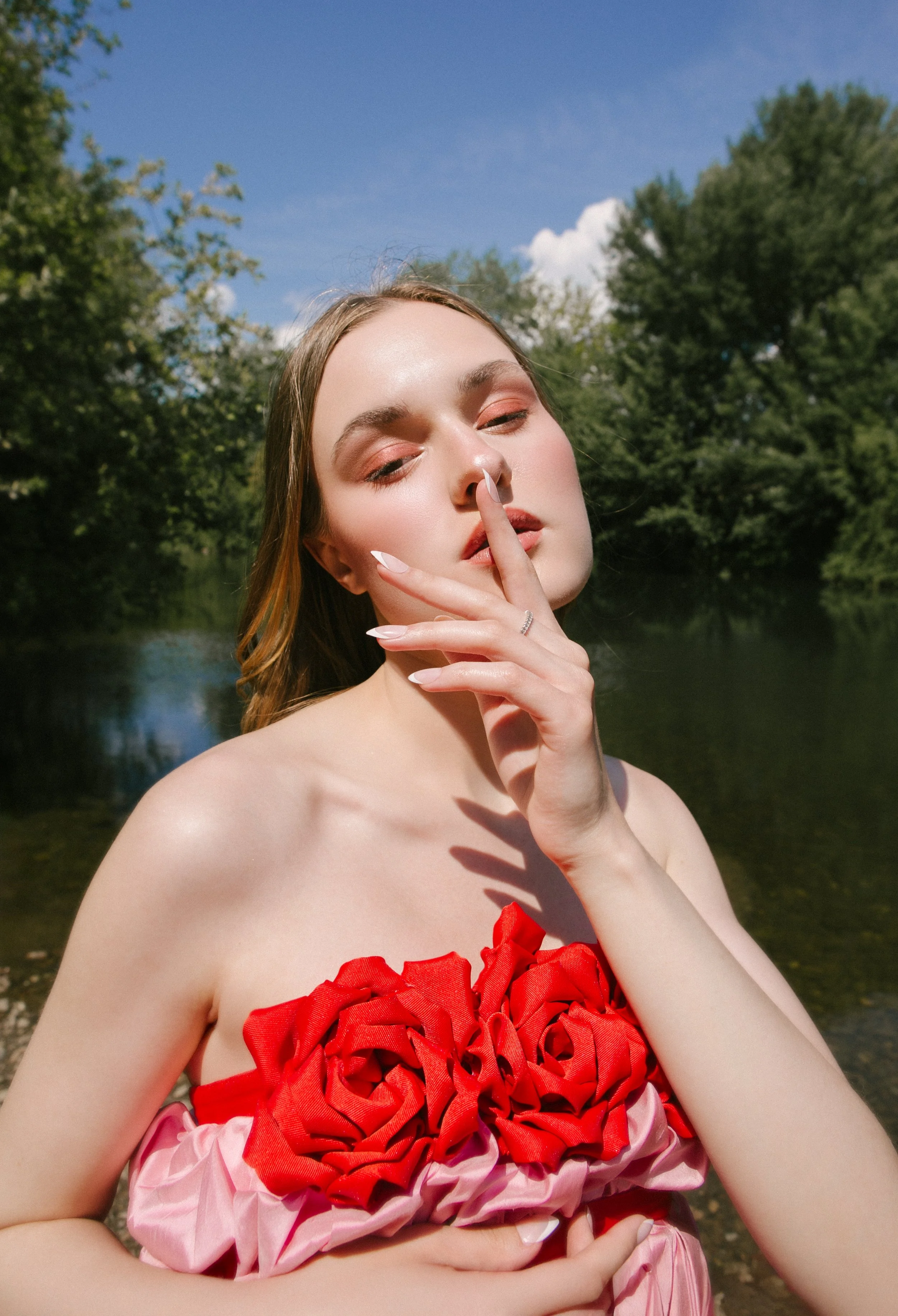 A young woman with fair skin and long hair stands outdoors near a body of water, wearing a strapless dress with red and pink fabric. She has her eyes closed and is touching her nose with her finger.