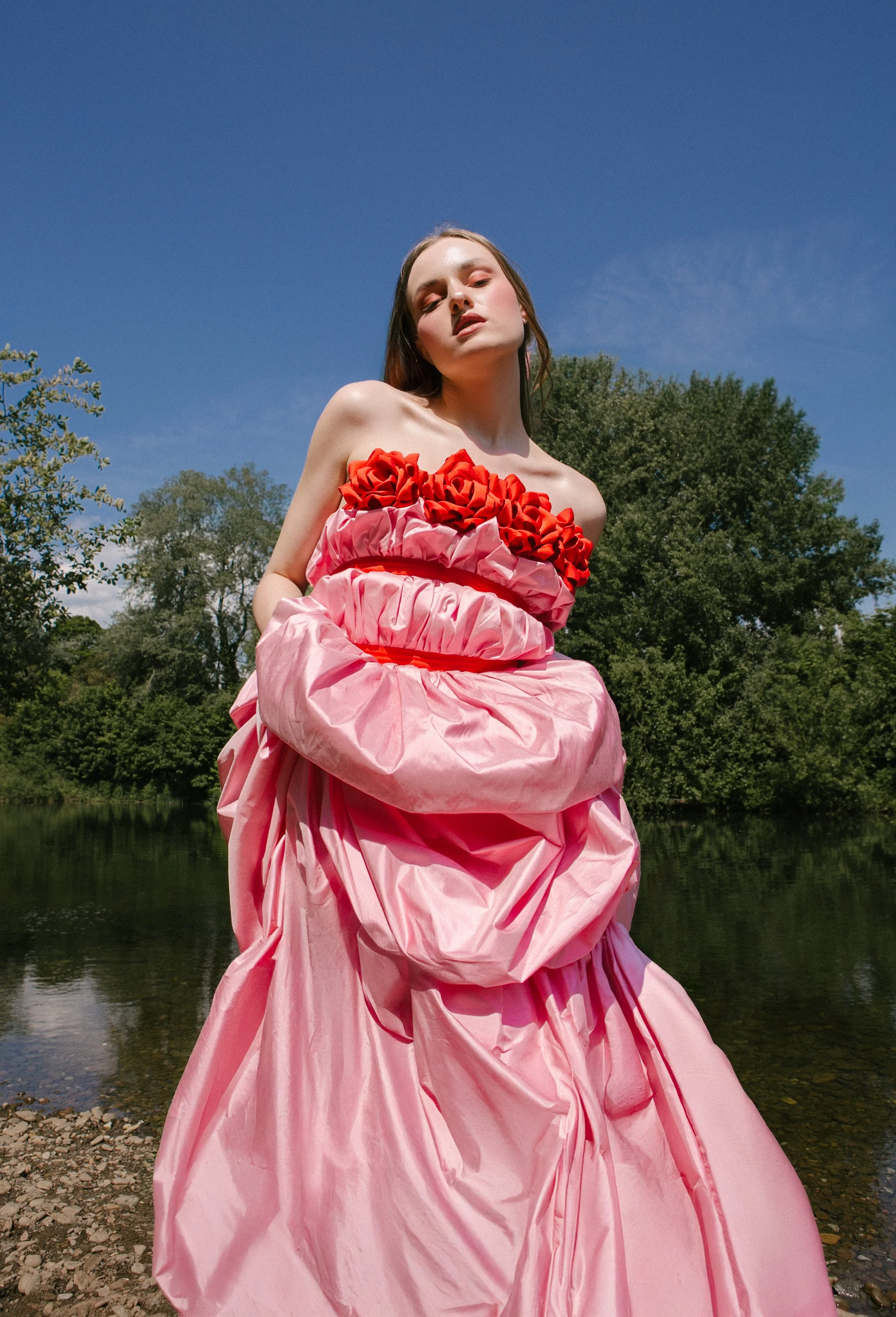 A woman wearing a pink dress with red and pink floral embellishments stands outdoors near a body of water with green trees and a blue sky in the background.