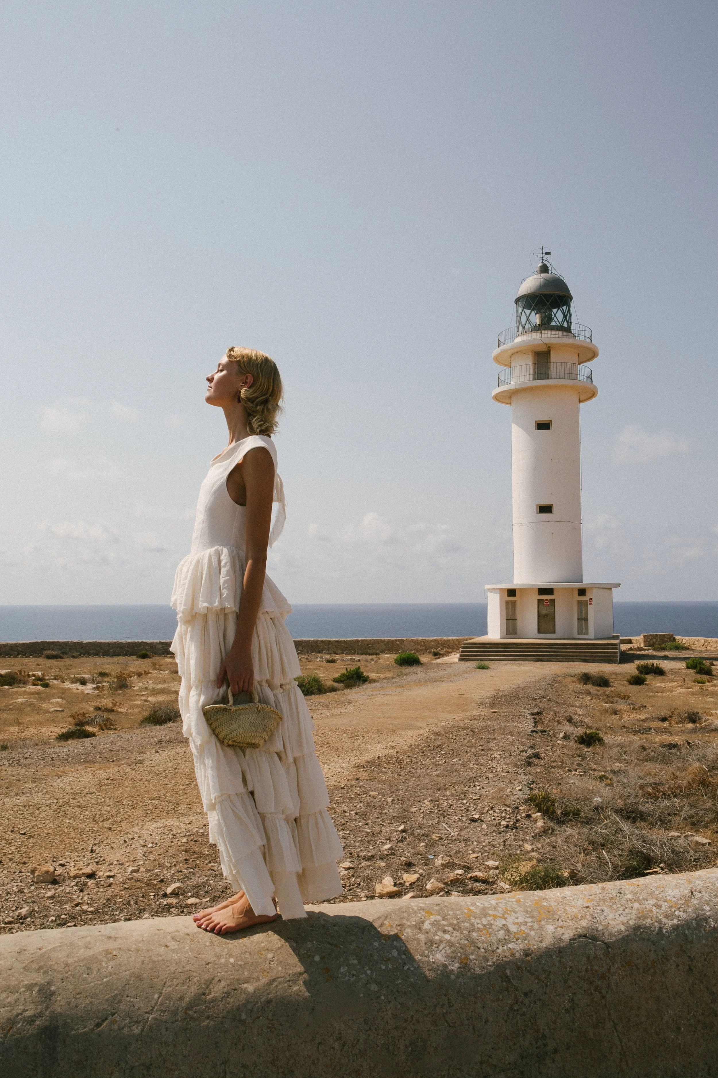 A woman in a white ruffled dress standing barefoot on a concrete ledge with her eyes closed near a lighthouse on a coastal landscape.