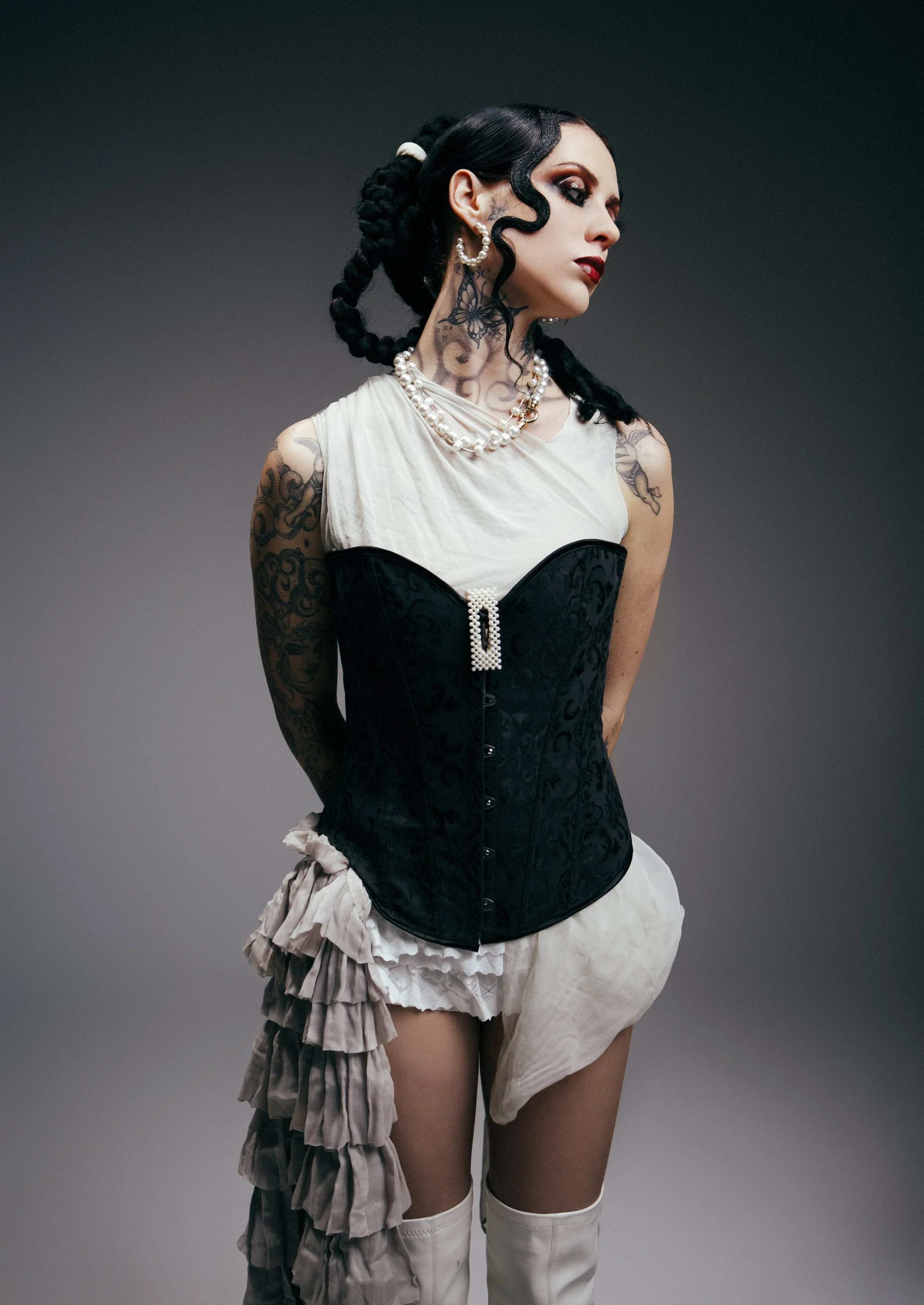 A woman with black hair styled in braids, tattoos on her arms and neck, wearing a white top, a black corset, layered ruffled skirt, thigh-high white boots, and jewelry including pearls and earrings, standing against a gray background.
