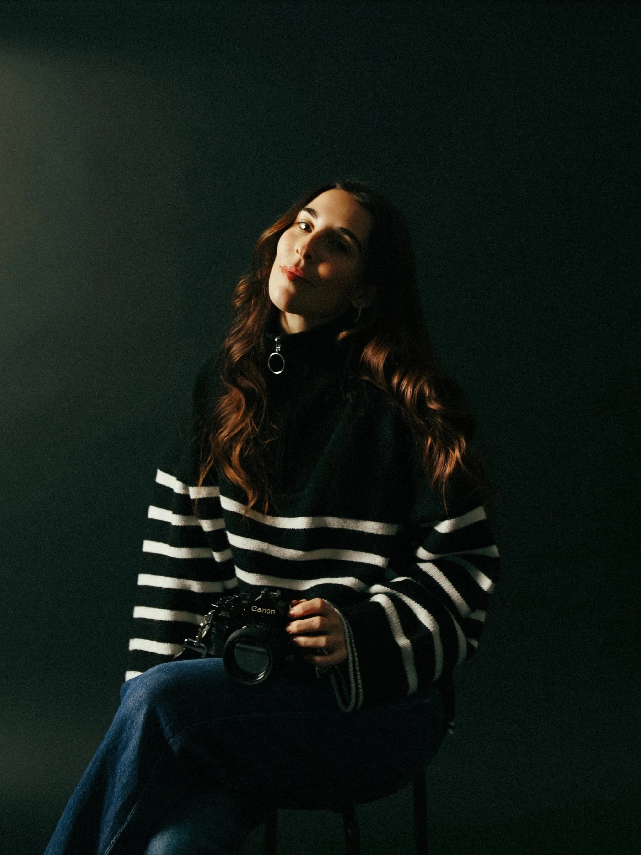 A young woman with long wavy hair sits against a dark background, holding a Canon camera. She wears a black and white striped sweater and looks at the camera with a slight smile.