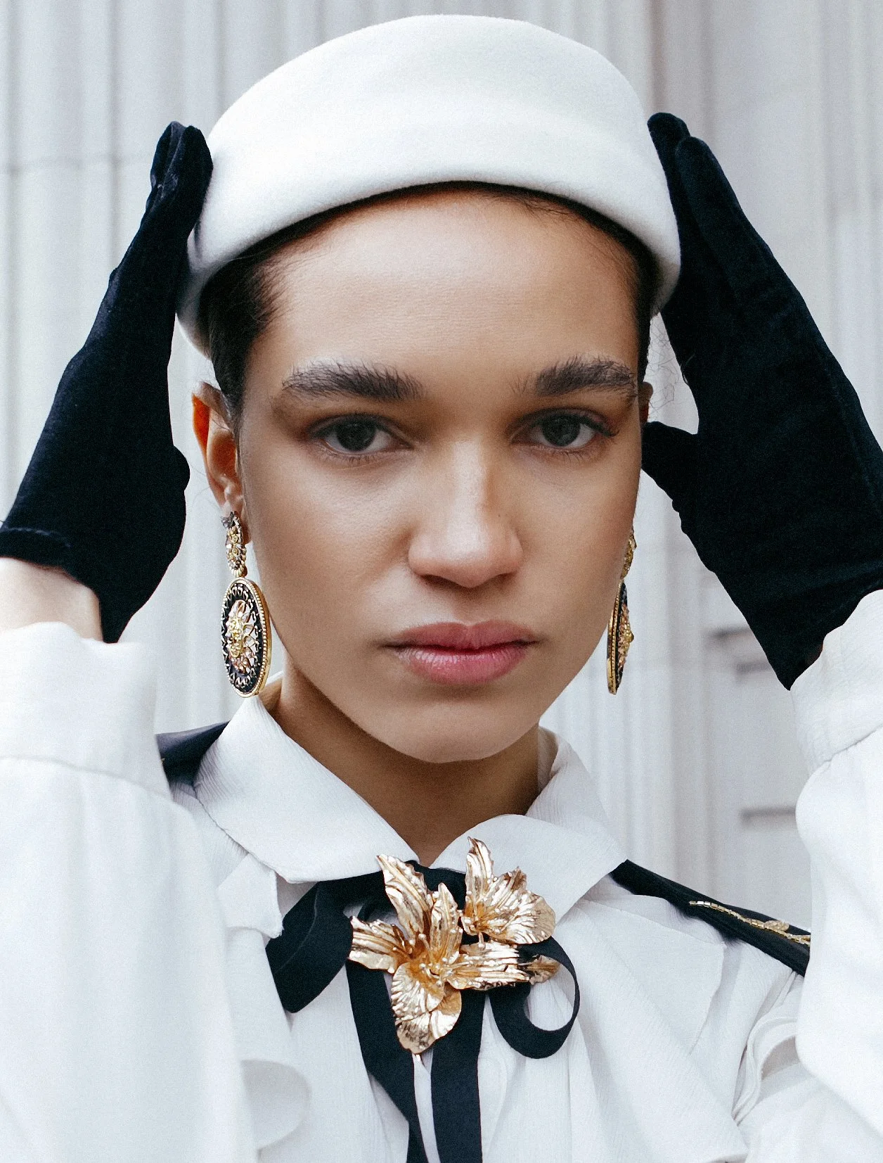 Close-up of a woman wearing a white hat, black gloves, ornate earrings, and a white blouse with a gold floral brooch.