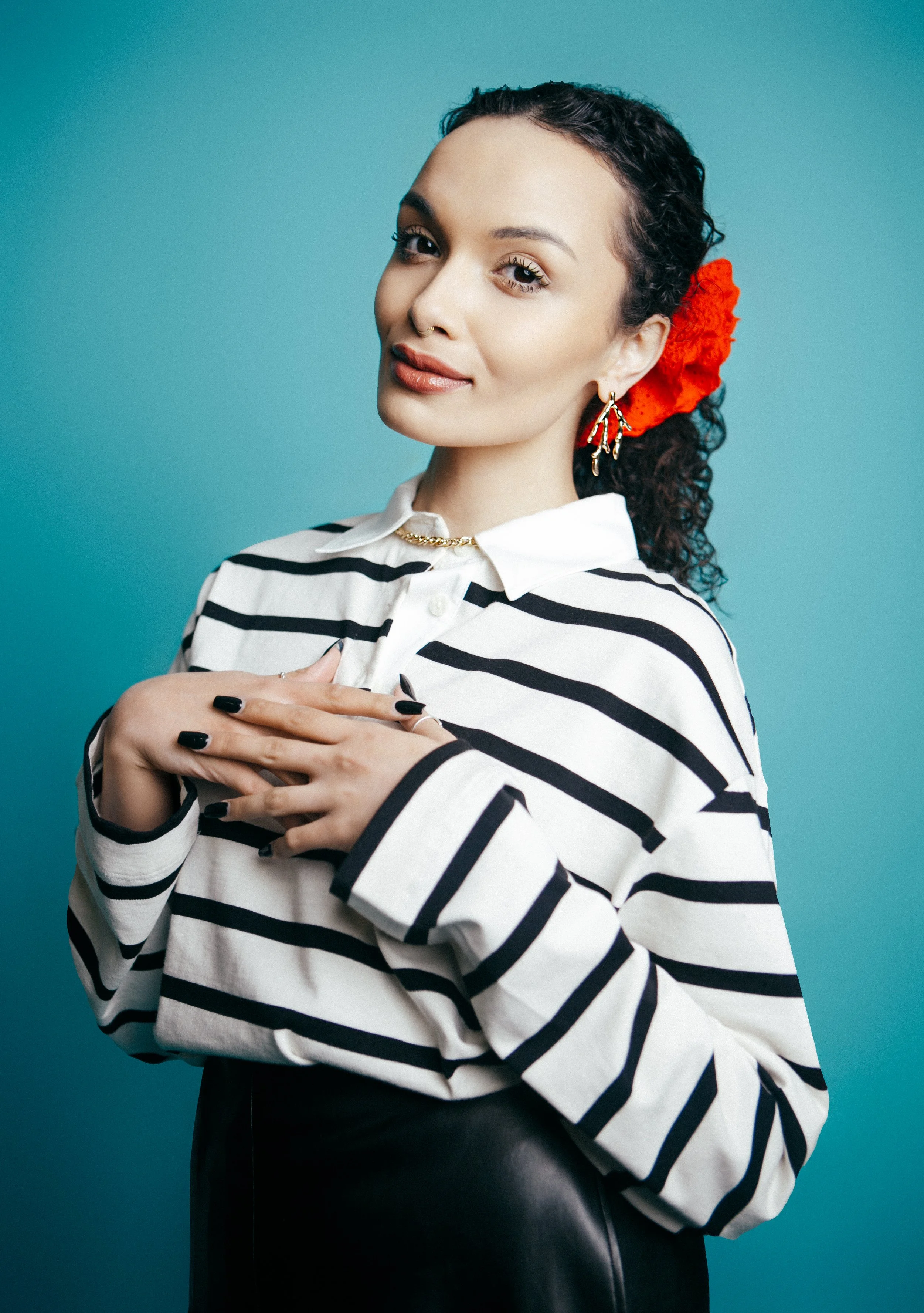 A young woman with curly dark hair, red hair accessory, and earrings. She wears a black and white striped blouse and has her hands crossed over her chest, standing against a turquoise background.