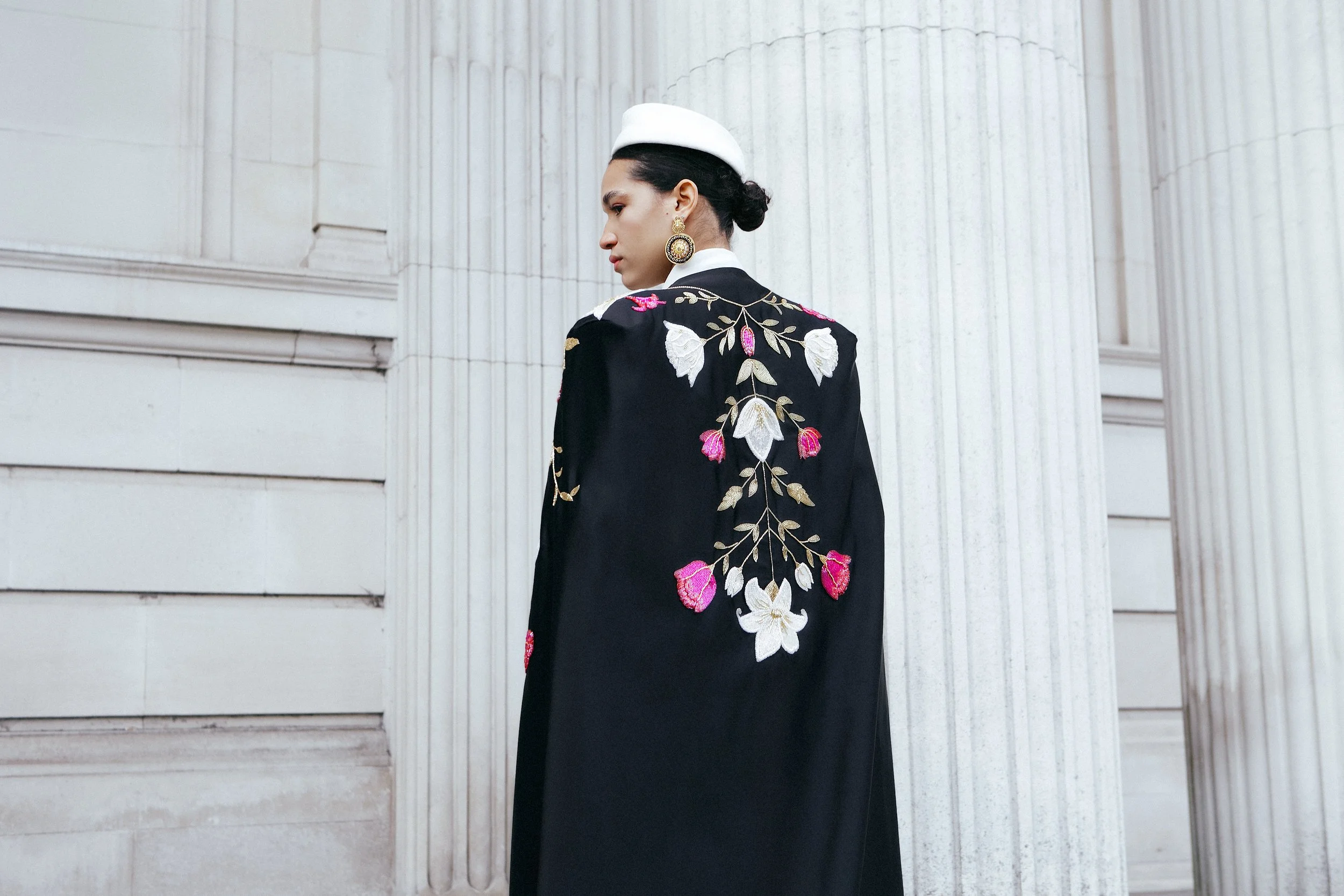 Woman in a black embroidered coat with floral design, white hat, and gold earrings standing against a white columned building.