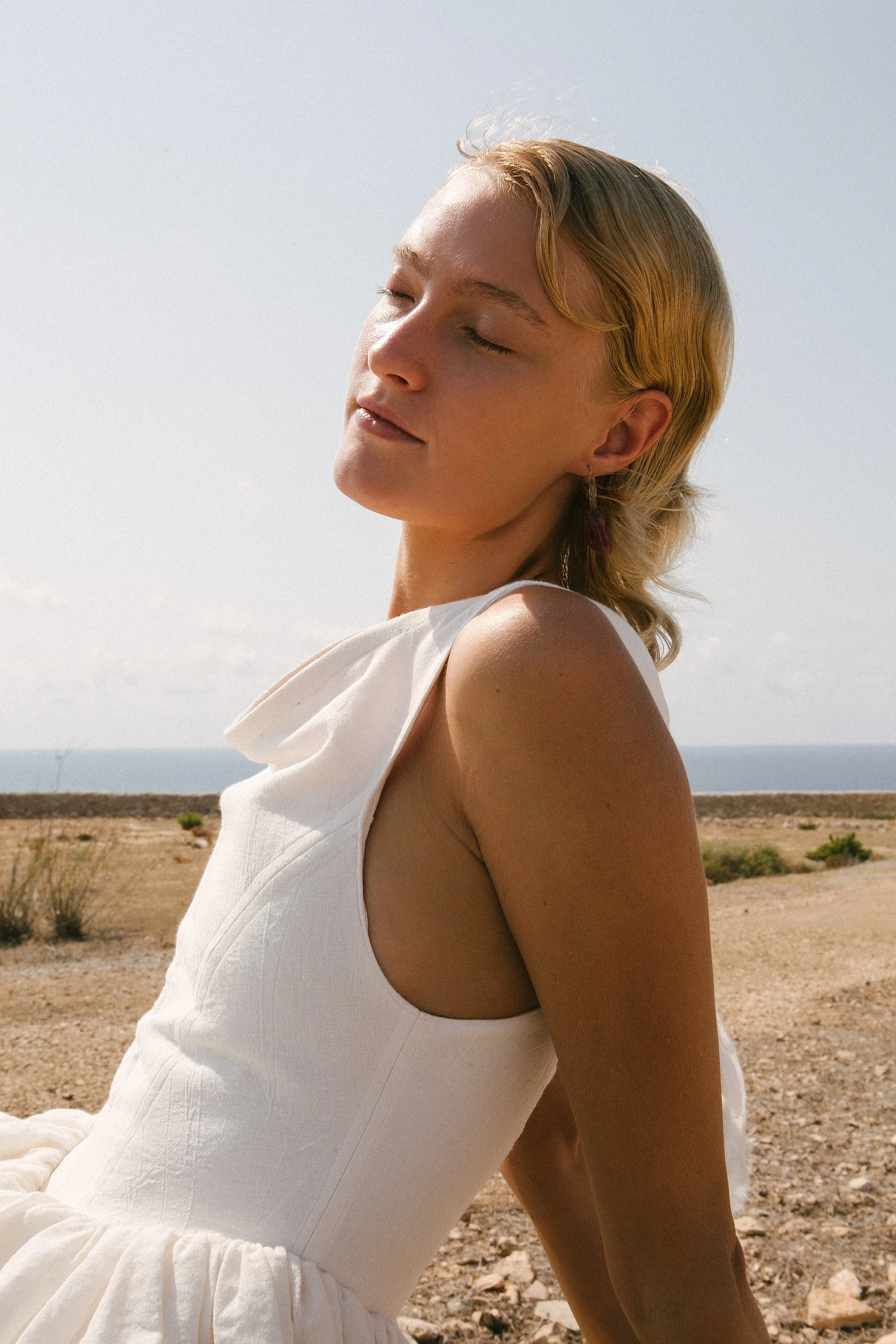 A woman with blonde hair and earrings sitting outdoors with her eyes closed, wearing a white sleeveless dress, in a dry, desert-like landscape with the ocean in the background.