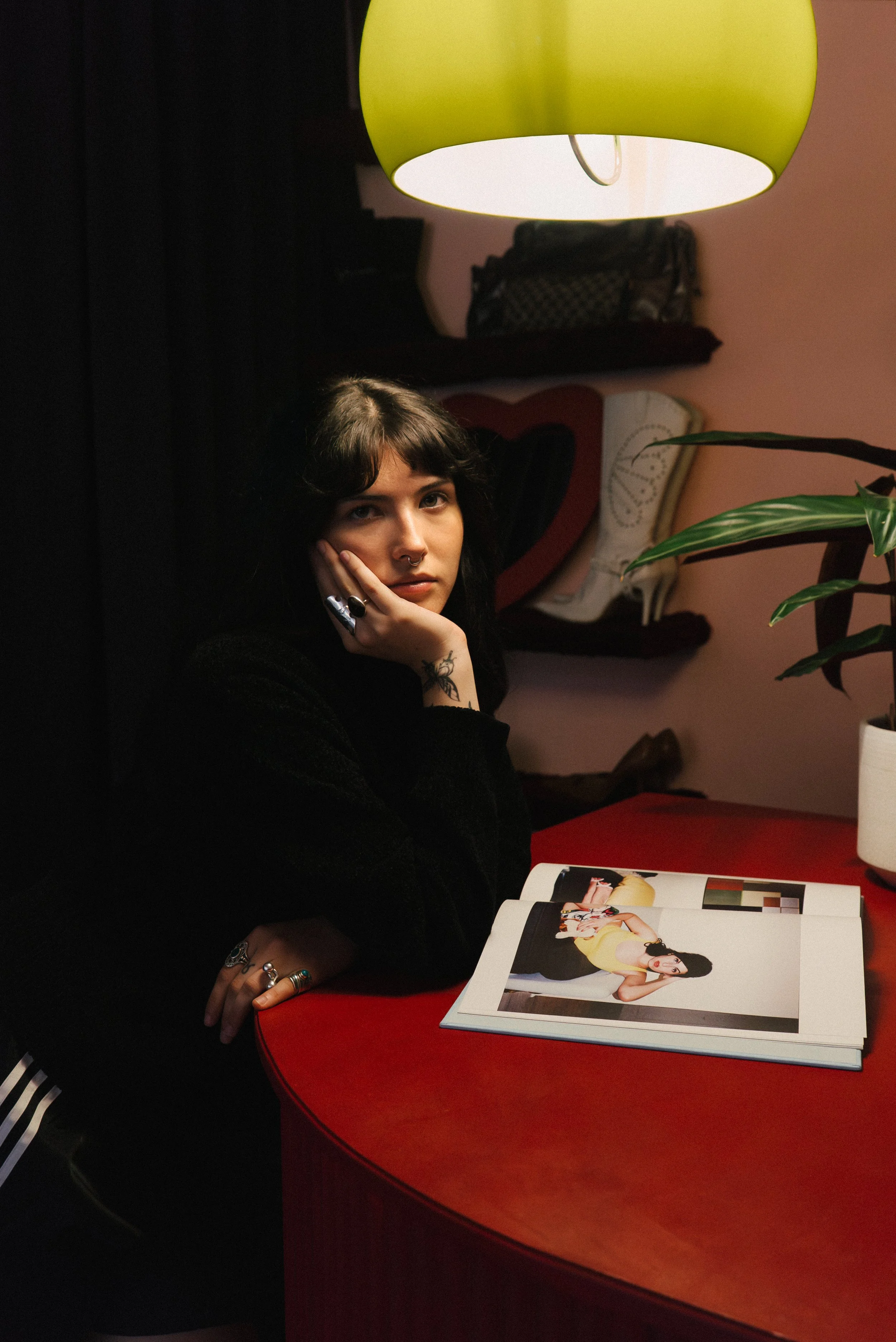 Young woman with dark hair, sitting at a red table, looking at the camera, with her hand resting on her cheek, in a dimly lit room with a green hanging lamp, an open magazine, a potted plant, and art on the wall behind her.
