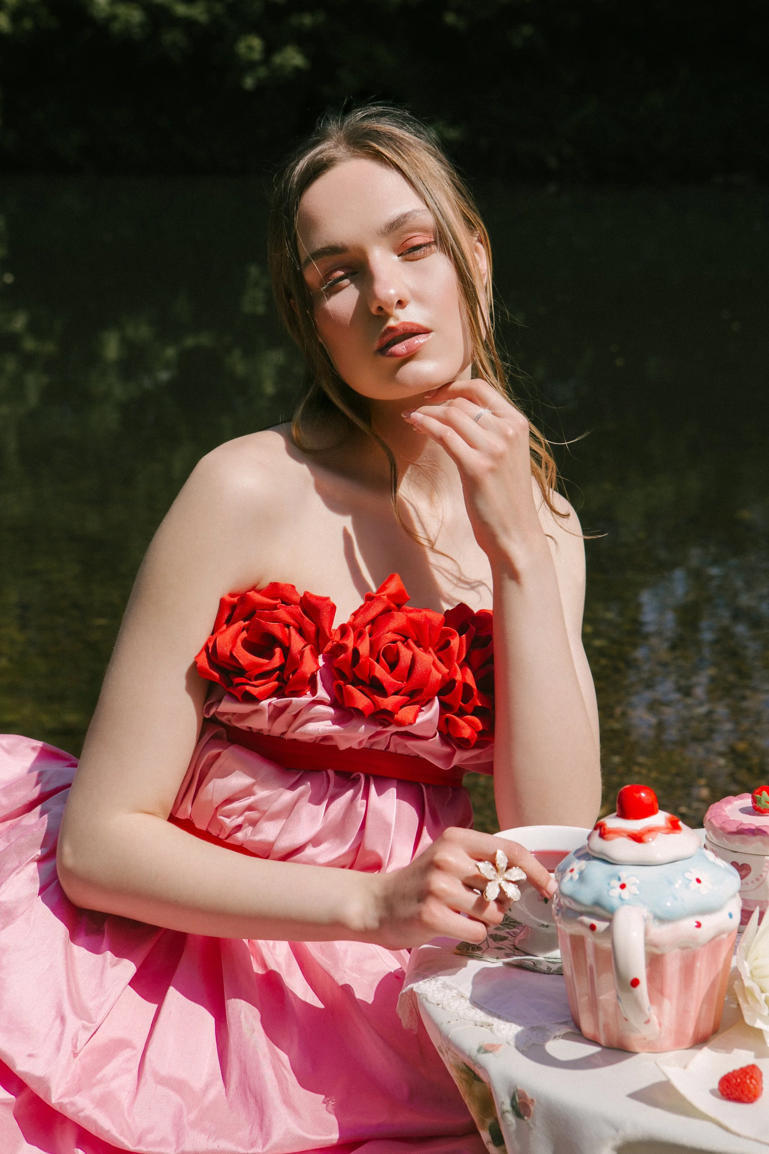A woman wearing a pink dress with red fabric roses on the top sits outdoors at a table with a cupcake decorated with a strawberry and whipped cream, surrounded by tea cups and plates.