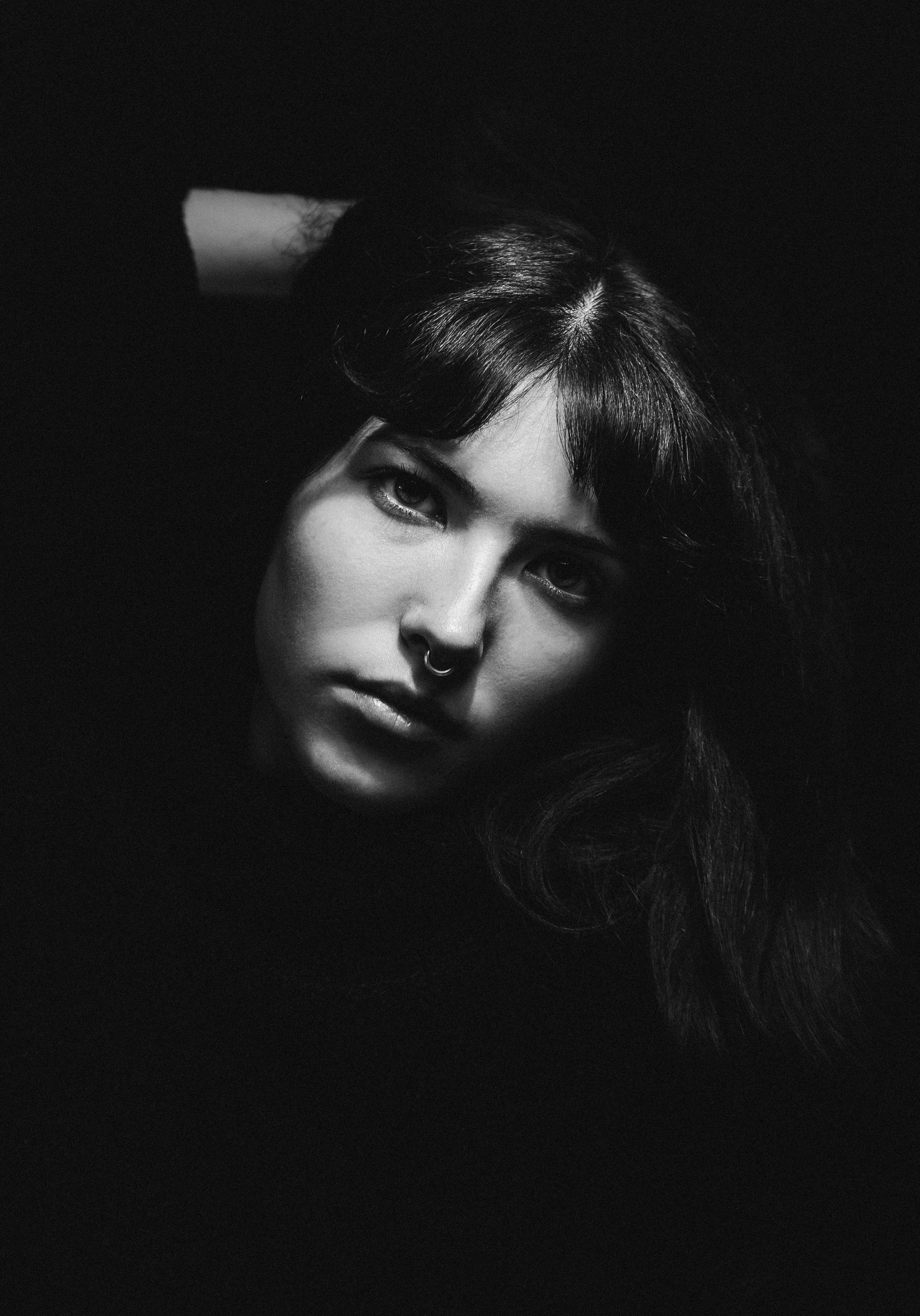 A black and white portrait of a young woman with dark hair, wearing a septum piercing, looking directly at the camera with a serious expression, against a dark background.