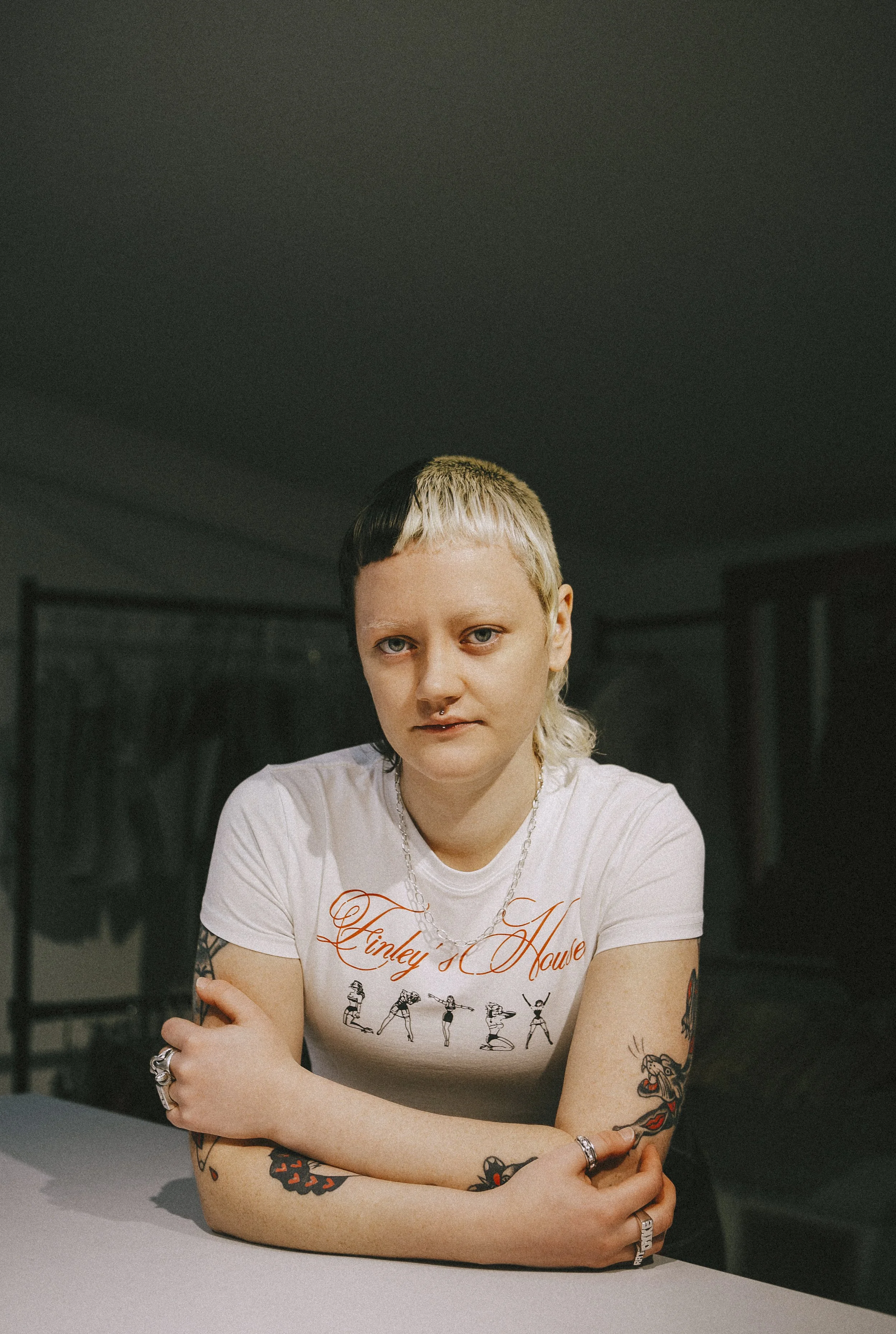 A young woman with short, two-tone hair (black and blonde) sitting at a table with her arms crossed, wearing a white t-shirt with the red script reading 'Hinley s House' and illustrations of women dancing. She has multiple tattoos on her arms and rings on her fingers, against a dark background.