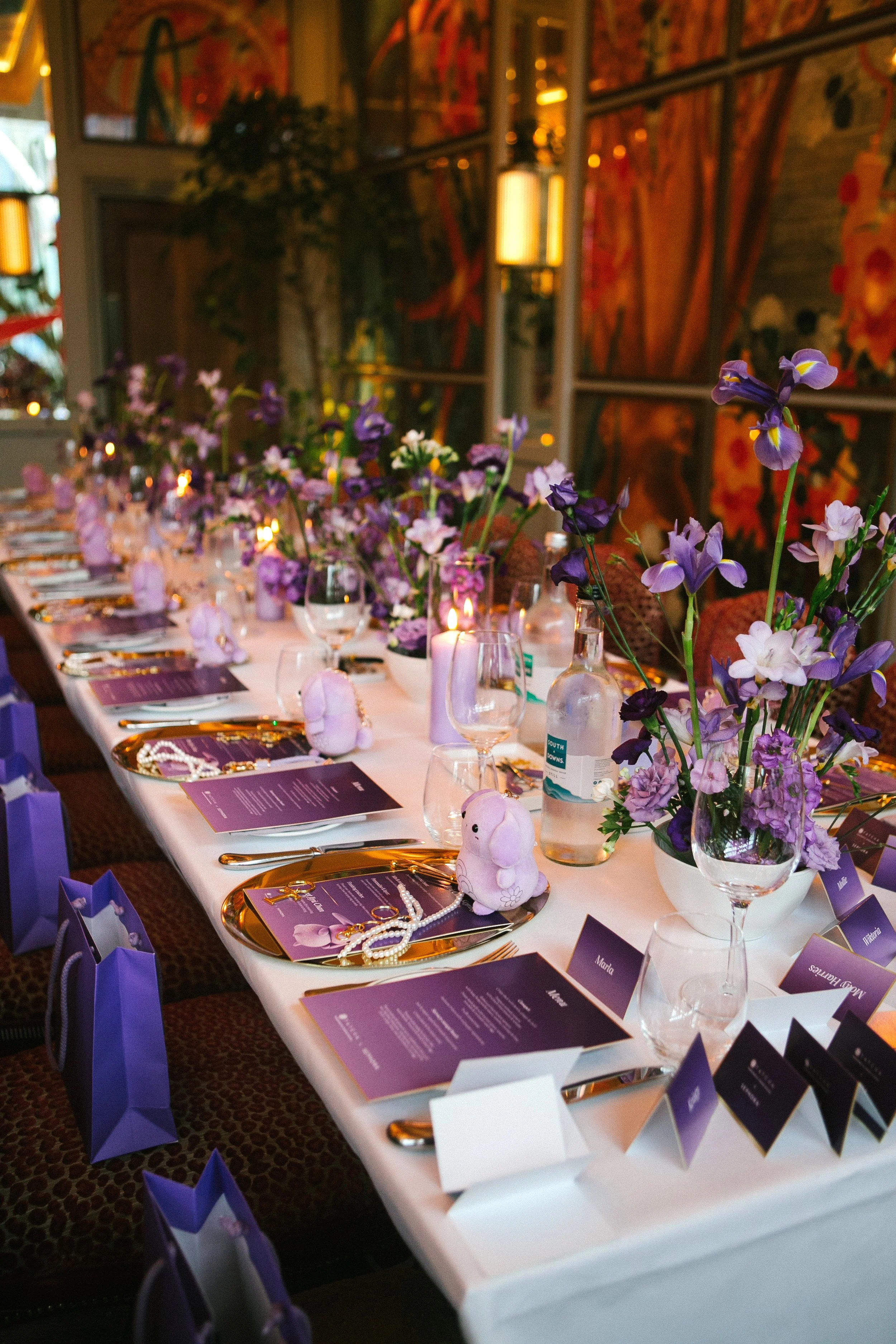 Decorated banquet table with purple flowers, place cards, glassware, gold utensils, and purple gift bags, set for a celebration or event in a warmly lit indoor venue.