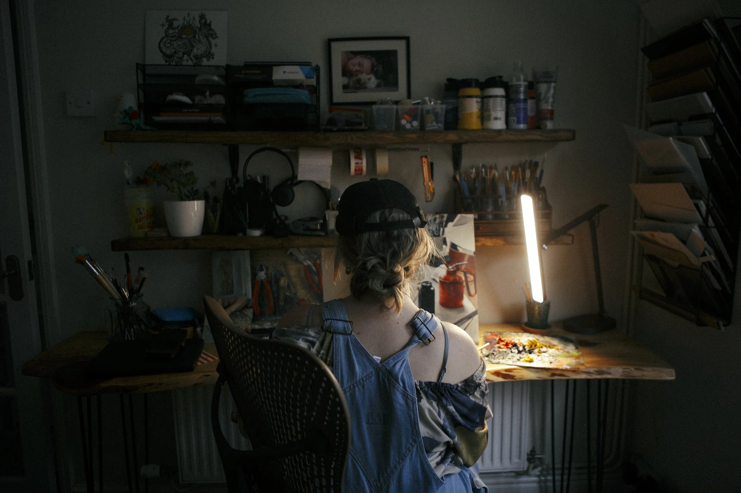 Person with curly hair, wearing a black cap and a sleeveless top, sitting at a wooden desk in a dimly lit art studio, working on a painting illuminated by a bright desk lamp.
