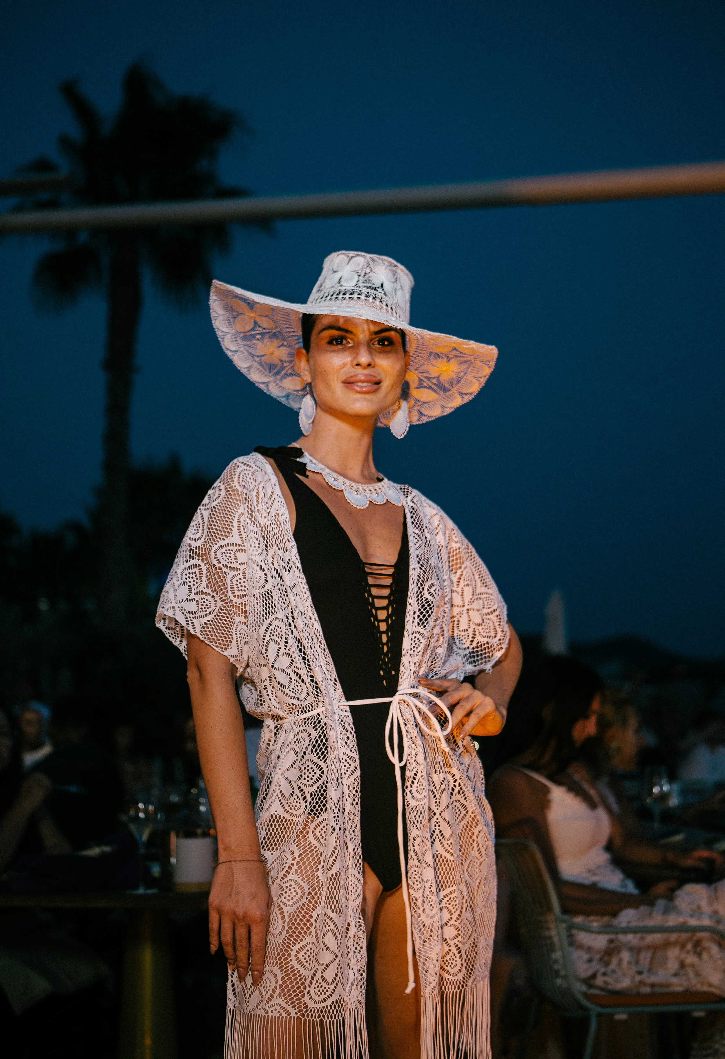 A woman in a wide-brimmed hat with lace detailing, wearing a black swimsuit with lace-up front, and a white lace cover-up with fringe, at an outdoor evening event.