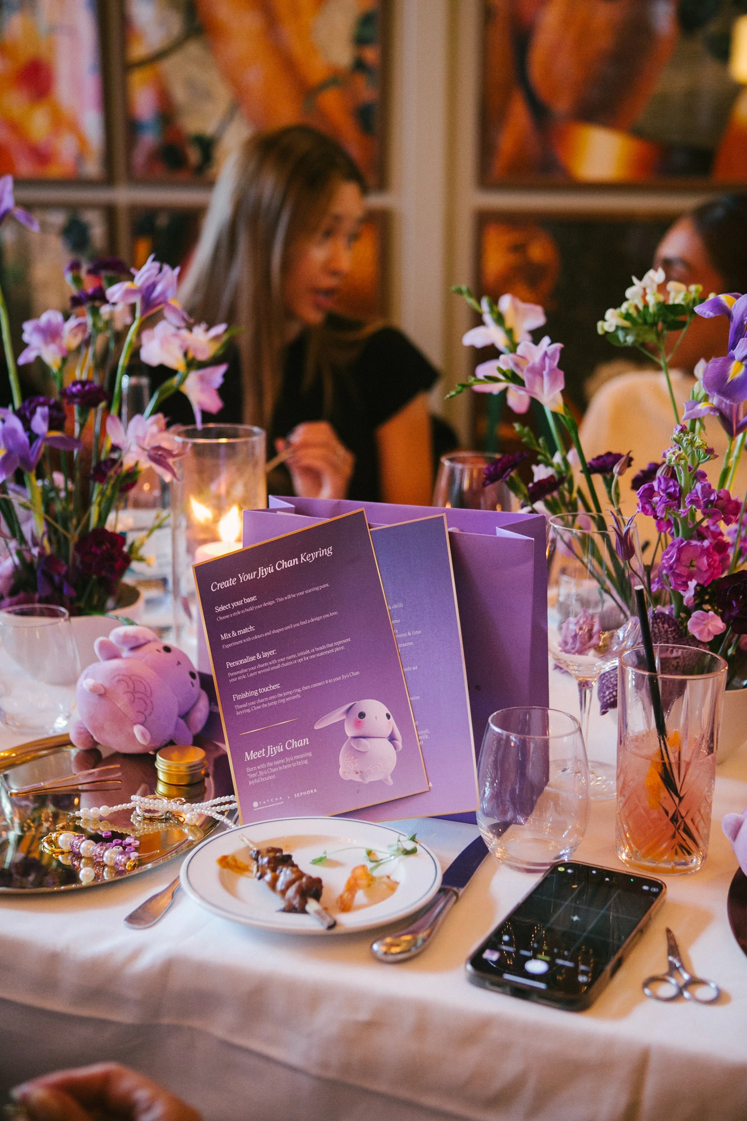 A decorated dining table with purple flowers, pink and purple drinks, and various tableware, including a smartphone, scissors, and jewelry. In the background, two women are blurred, seated at the table in a warmly lit room with colorful art on the wa