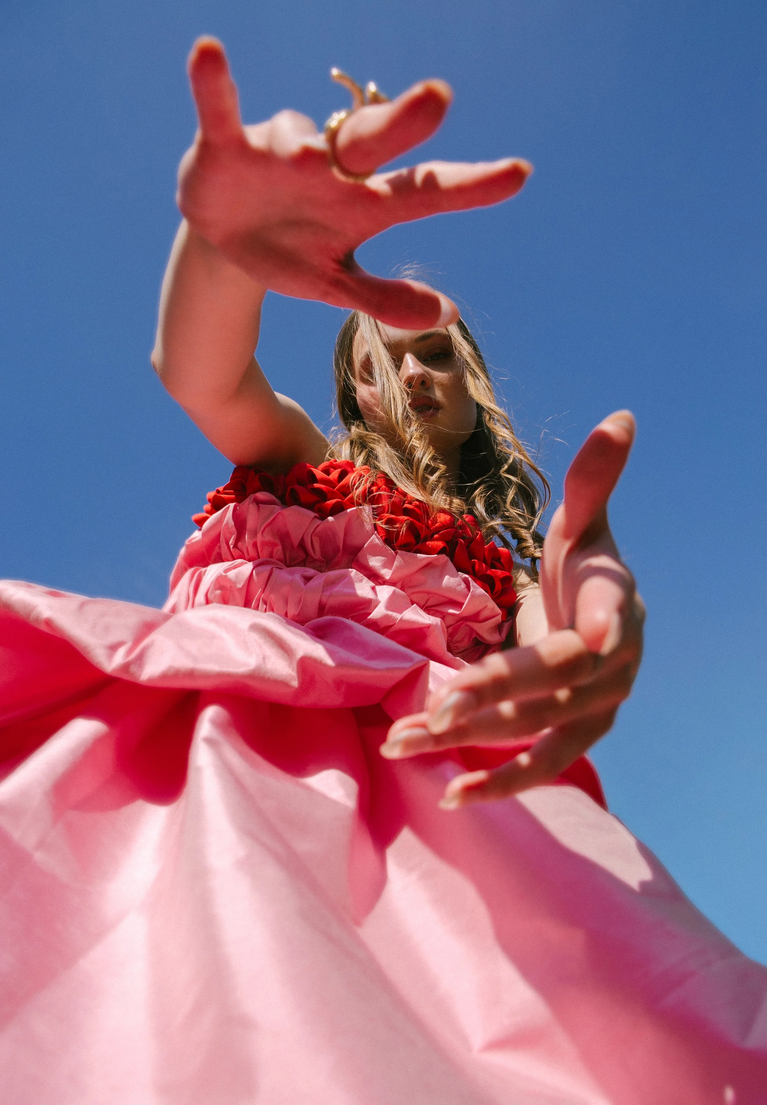 Low-angle shot of a woman in a pink dress with red decorative elements, reaching toward the camera against a clear blue sky.