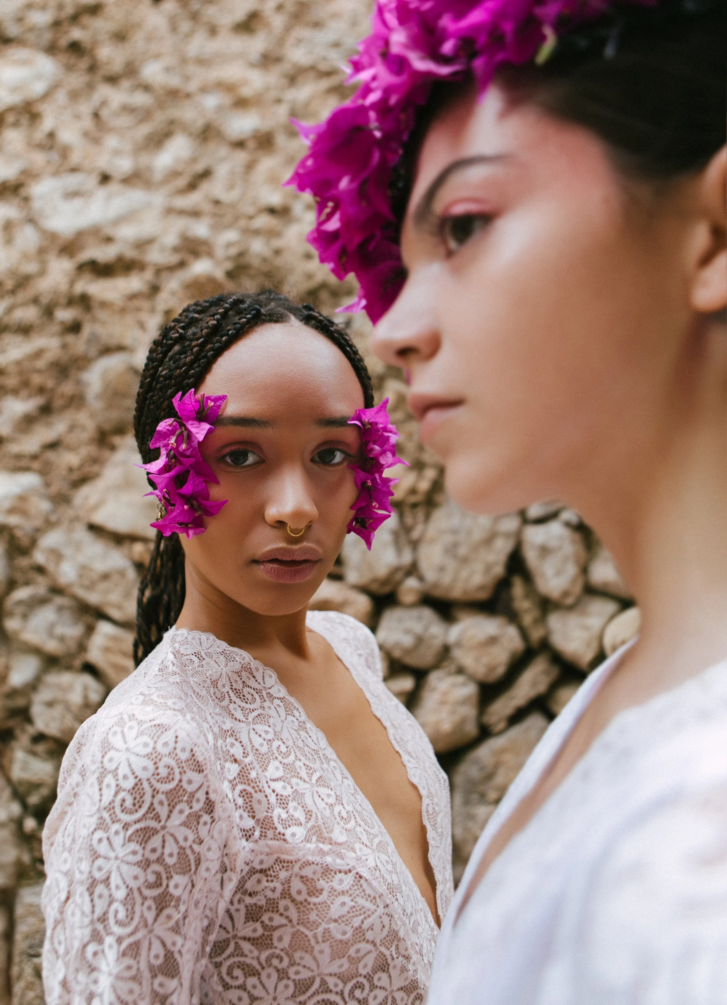 Two women with flowers on their faces and headpieces, one with braided hair and a septum piercing, in front of a stone wall.
